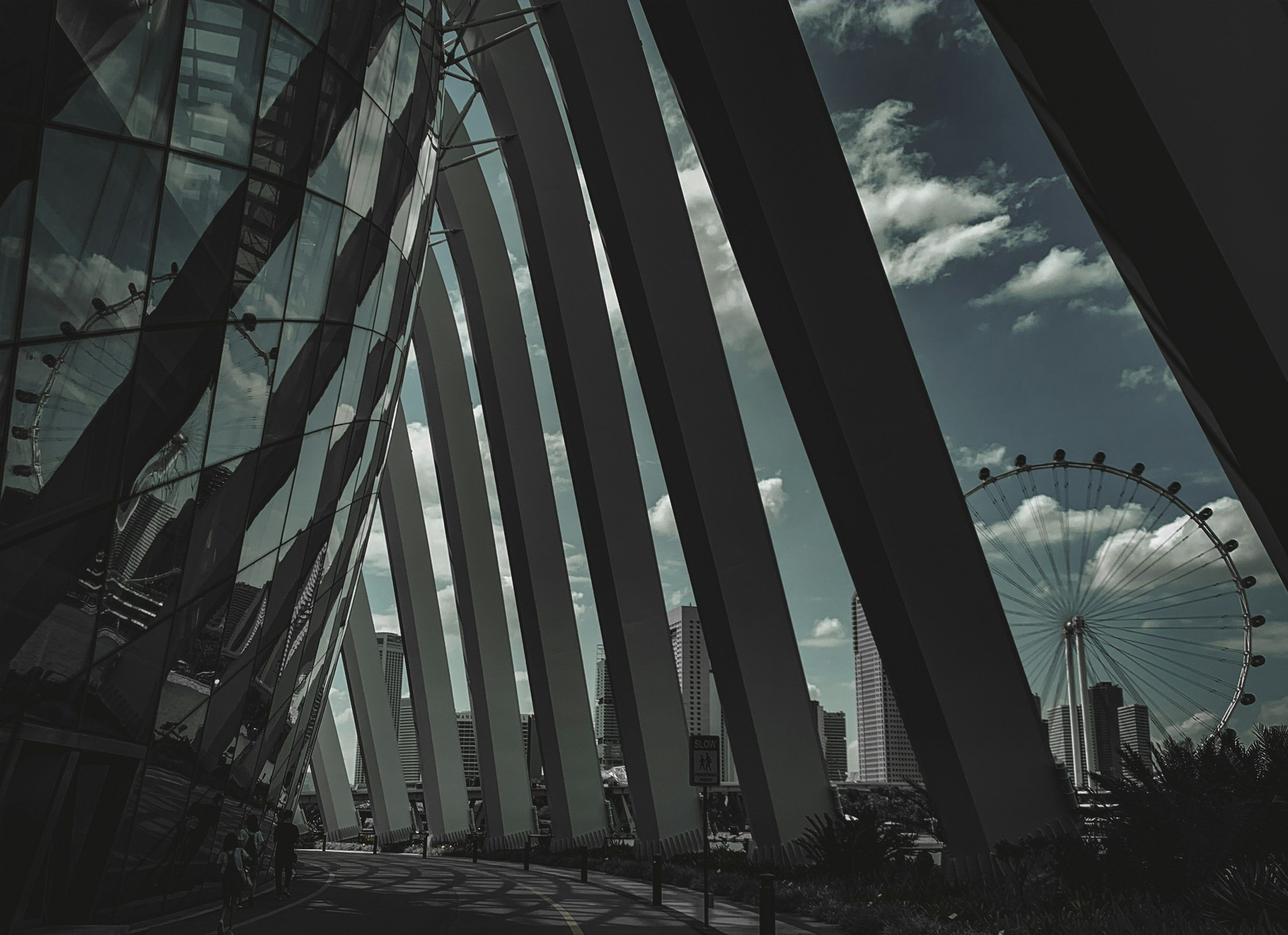 Modern architectural building with curved glass and steel design, city skyline with skyscrapers and a large Ferris wheel in the background, under a partly cloudy sky.