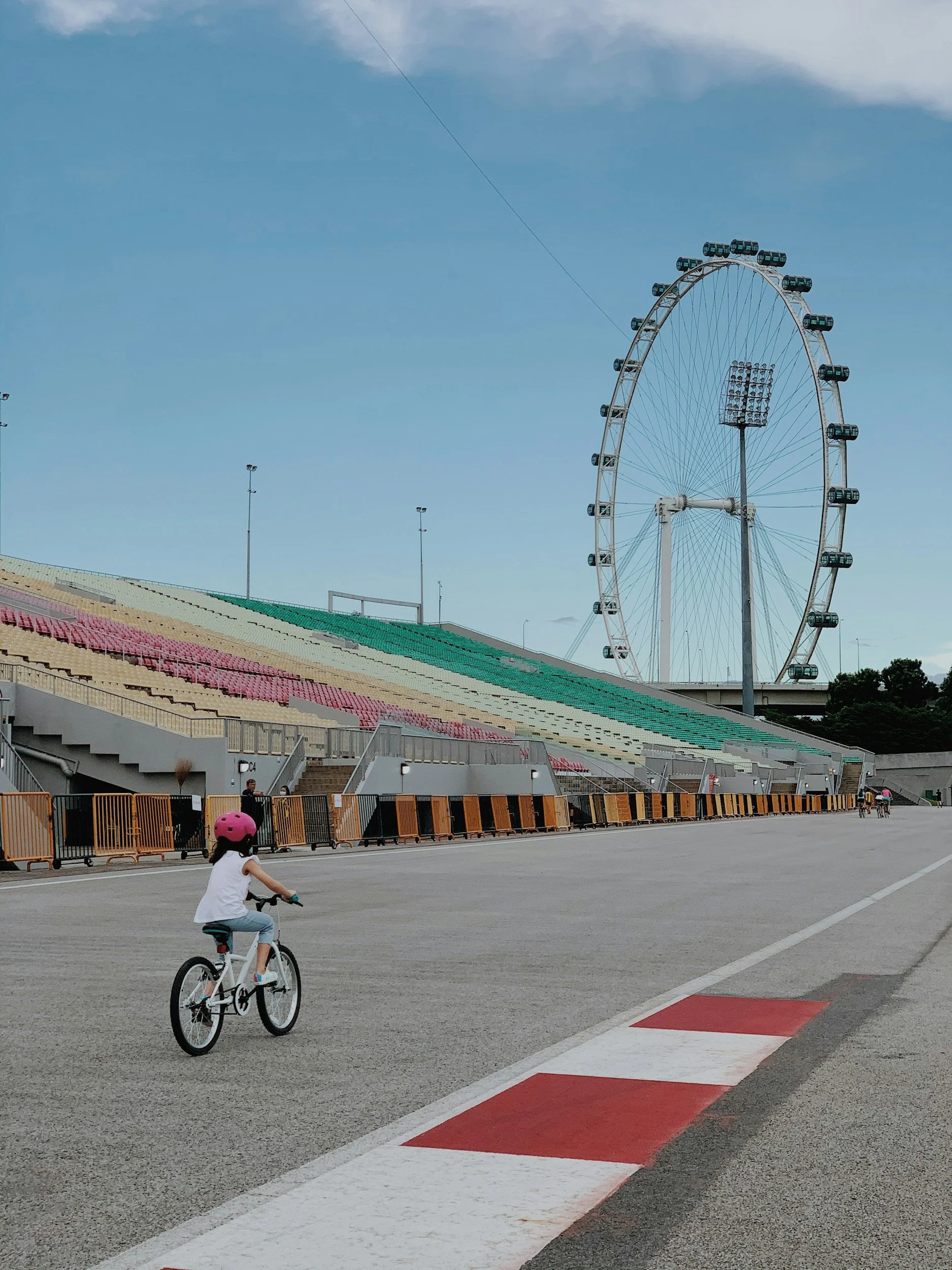 A young girl wearing a pink helmet riding a bicycle on a paved track with a large Ferris wheel in the background.