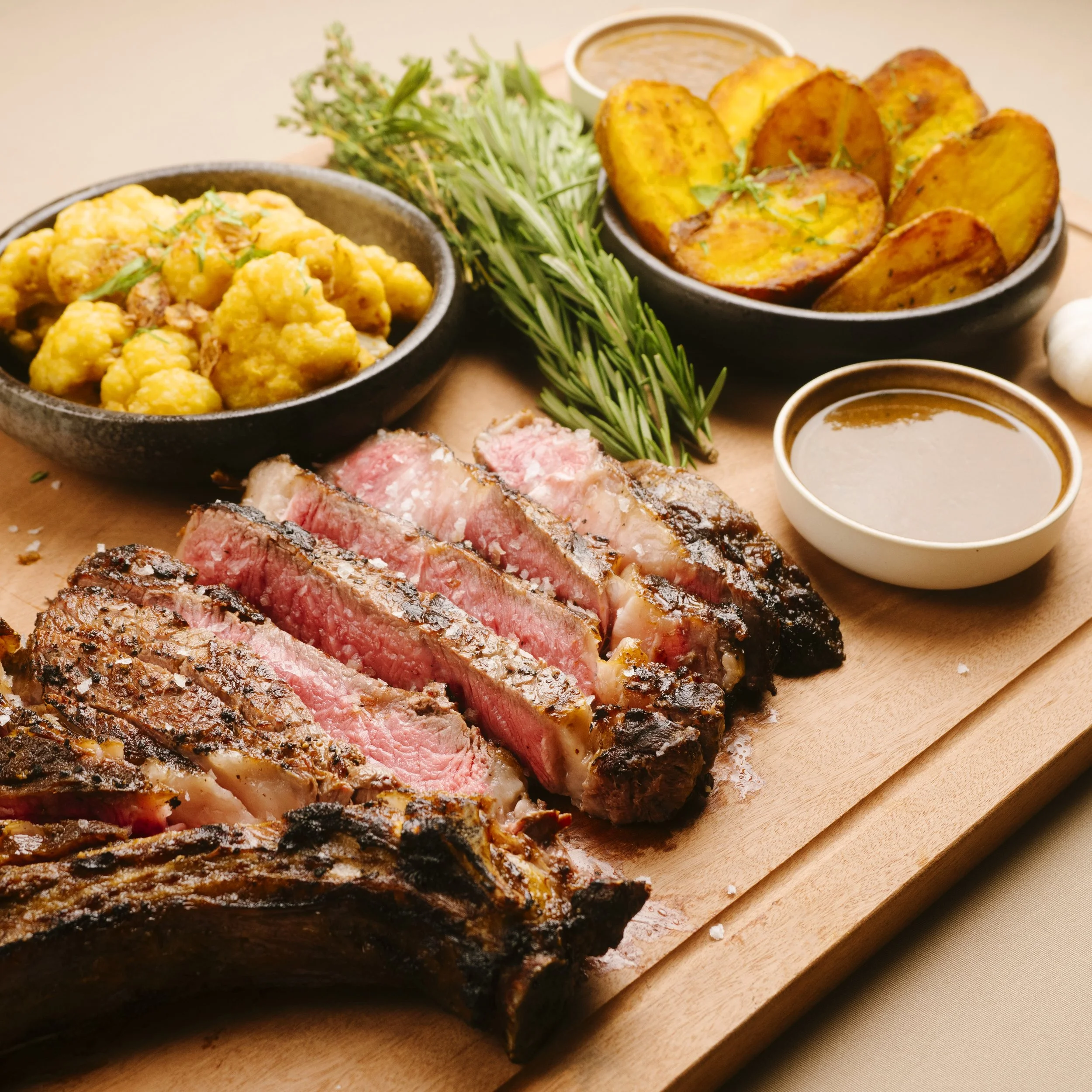 A plate of sliced medium-rare steak with roasted potatoes, cauliflower, fresh herbs, and dipping sauces on a wooden board.
