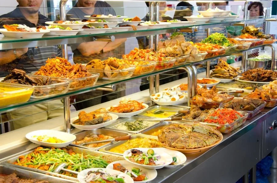 A buffet display with various dishes including salads, meats, baked goods, and vegetables, with people browsing in the background.