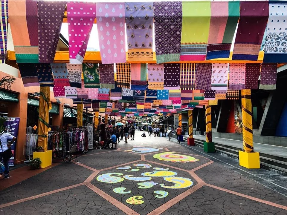 Colorful fabric banners hanging overhead in a marketplace street with people shopping beneath.