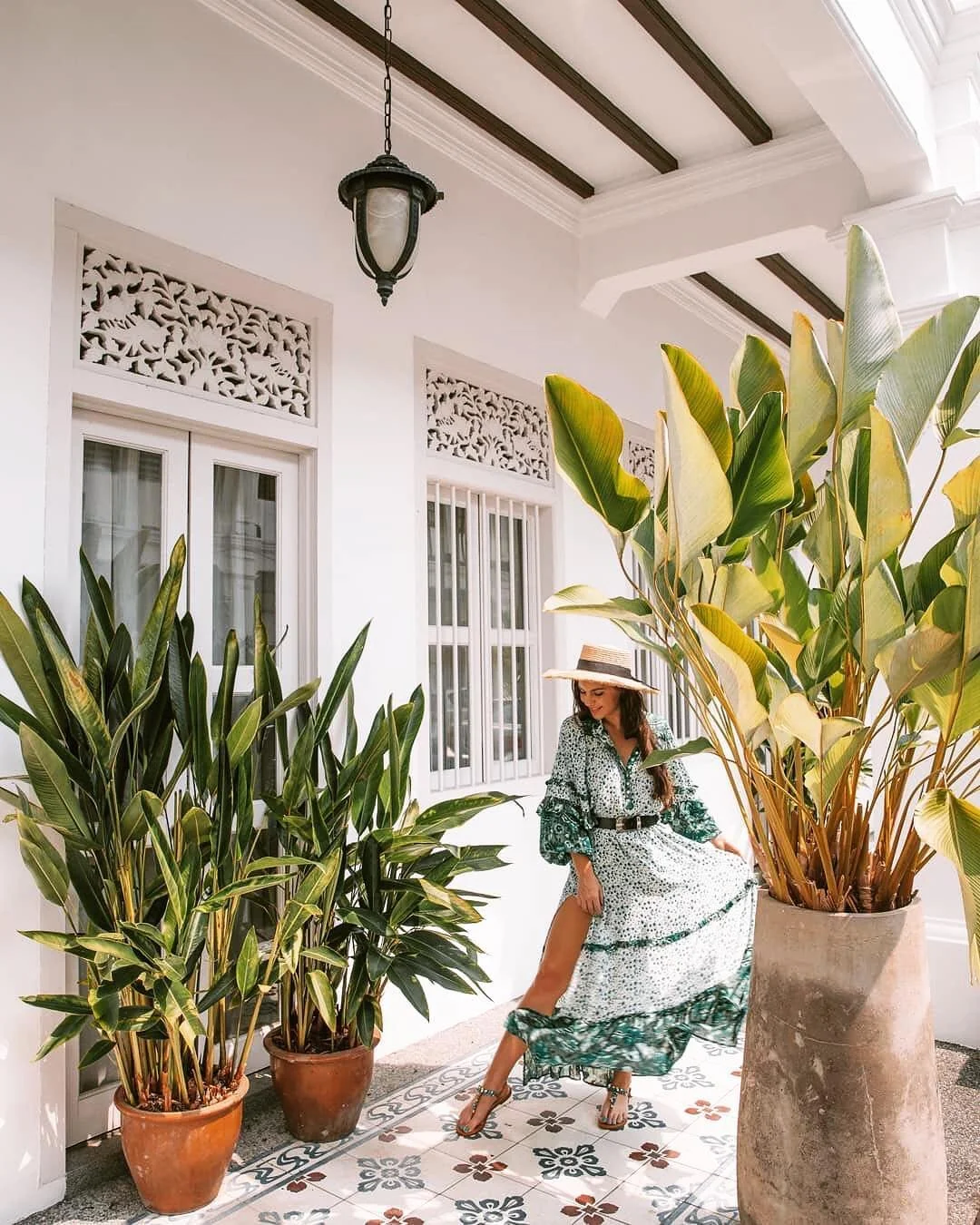 A woman in a flowing patterned dress with a slit, straw hat, and high heels stands on a patterned tile floor, surrounded by large potted tropical plants outside a white building with large windows and decorative woodwork.