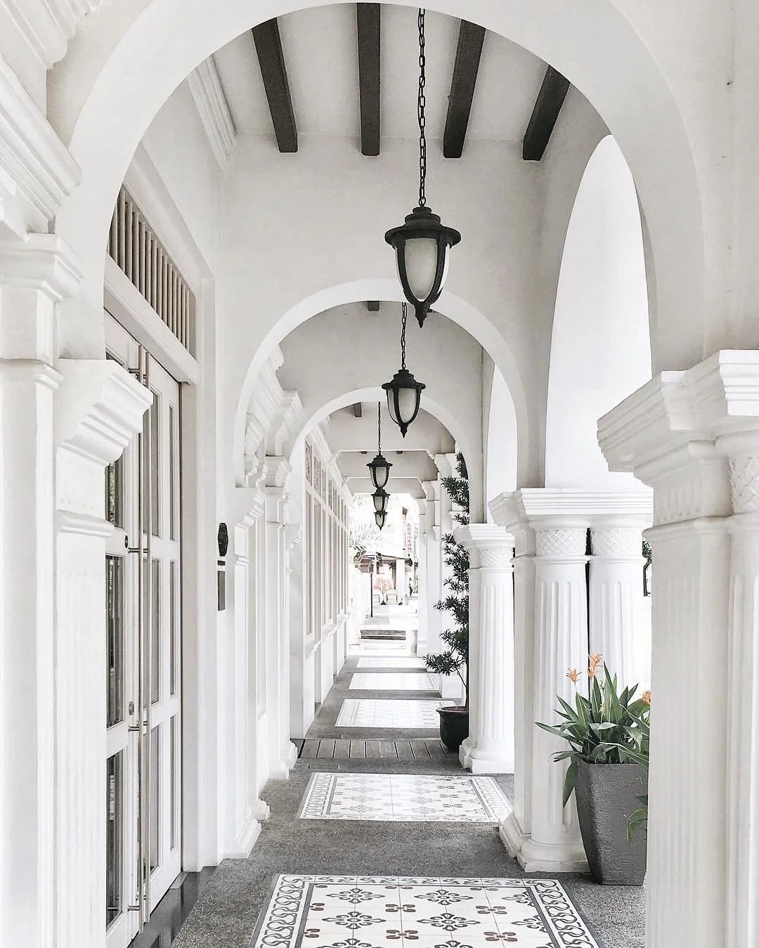 Covered porch with white walls, arches, black ceiling beams, hanging lantern lights, potted plants, and decorative tiled flooring.