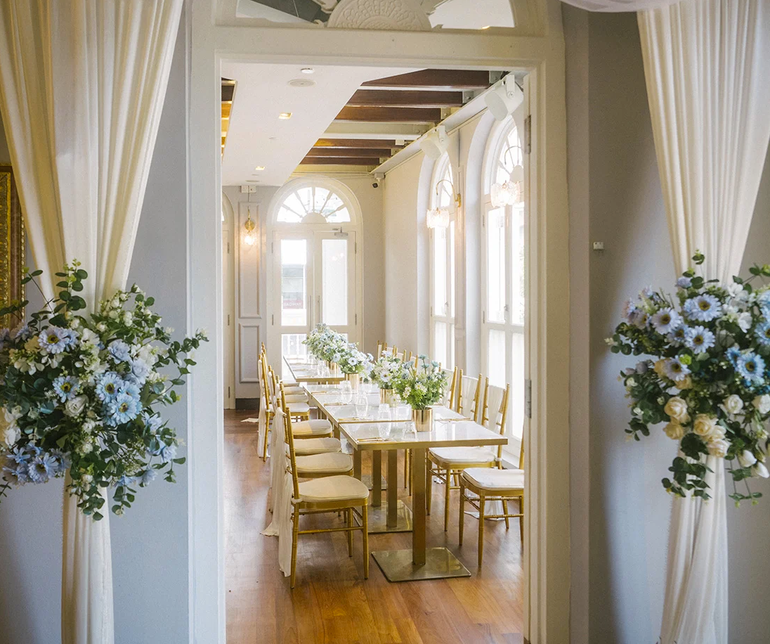 Elegant dining area with a long table arranged for a formal event, decorated with white flowers and gold chairs, surrounded by large windows and soft lighting.