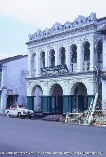 A two-story colonial-style building with arched windows and a balcony, featuring a sign that reads 'MUMBAI PRESS' at the front. There are cars parked in front and a person sitting nearby.