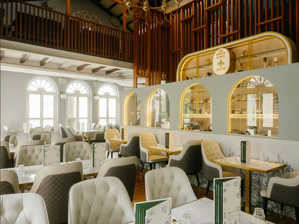 Elegant restaurant interior with white and beige tufted chairs, large arched windows, and decorative wall arches with gold accents, featuring a wooden balcony and shelving unit with candles and décor.