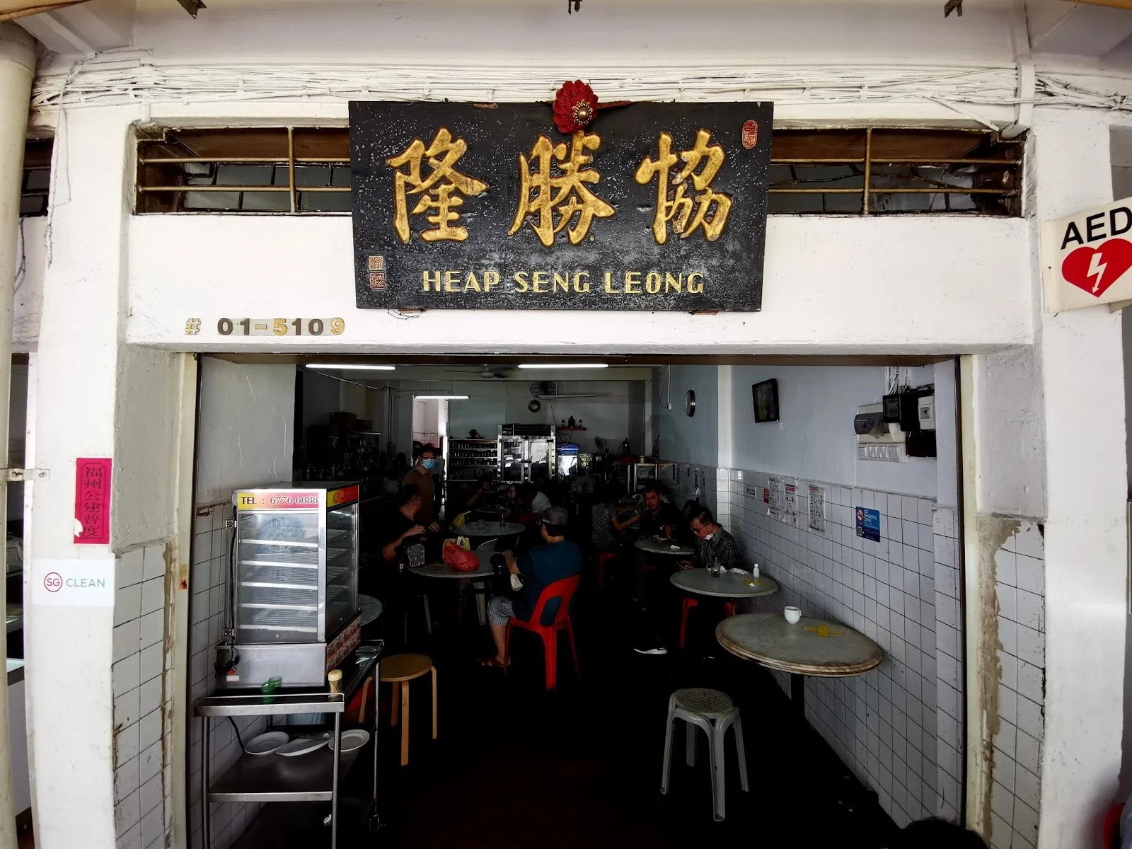 The entrance of a restaurant named Heap Seng Leong with a black signboard displaying Chinese characters and the name in English. Inside, there are several tables with people dining, some with bowls and drinks. The interior has white tiled walls and some electrical panels and notices on the side wall. An AED sign is visible outside the entrance.