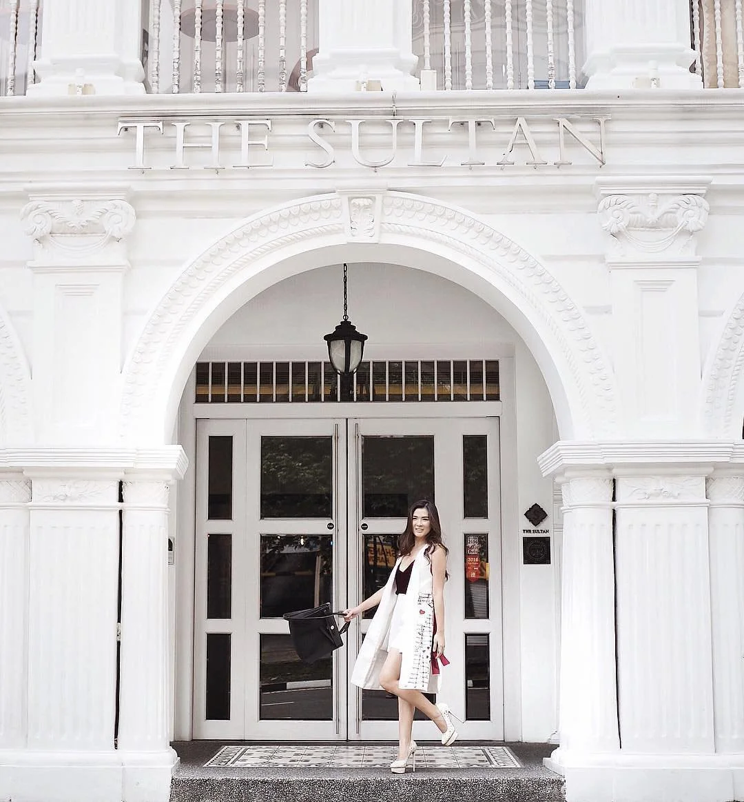 A woman standing in front of the entrance to The Sultan building, which has a white ornate facade with an archway, columns, and black door handles. She is smiling, holding a black bag, and wearing a white sleeveless dress with embroidery and a long vest.