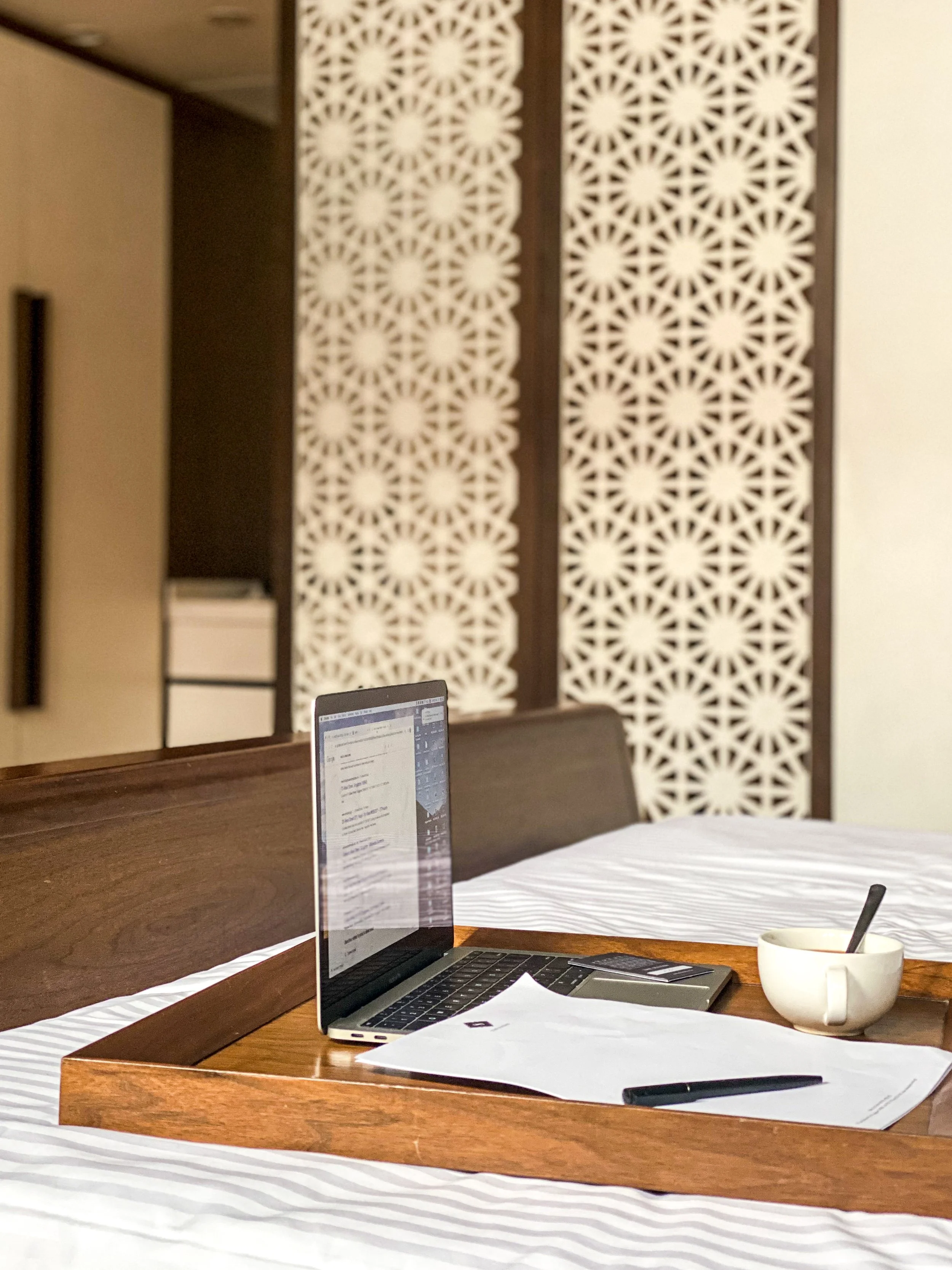 Laptop, papers, a pen, and a cup on a wooden tray on a bed, with decorative wall panels in the background.