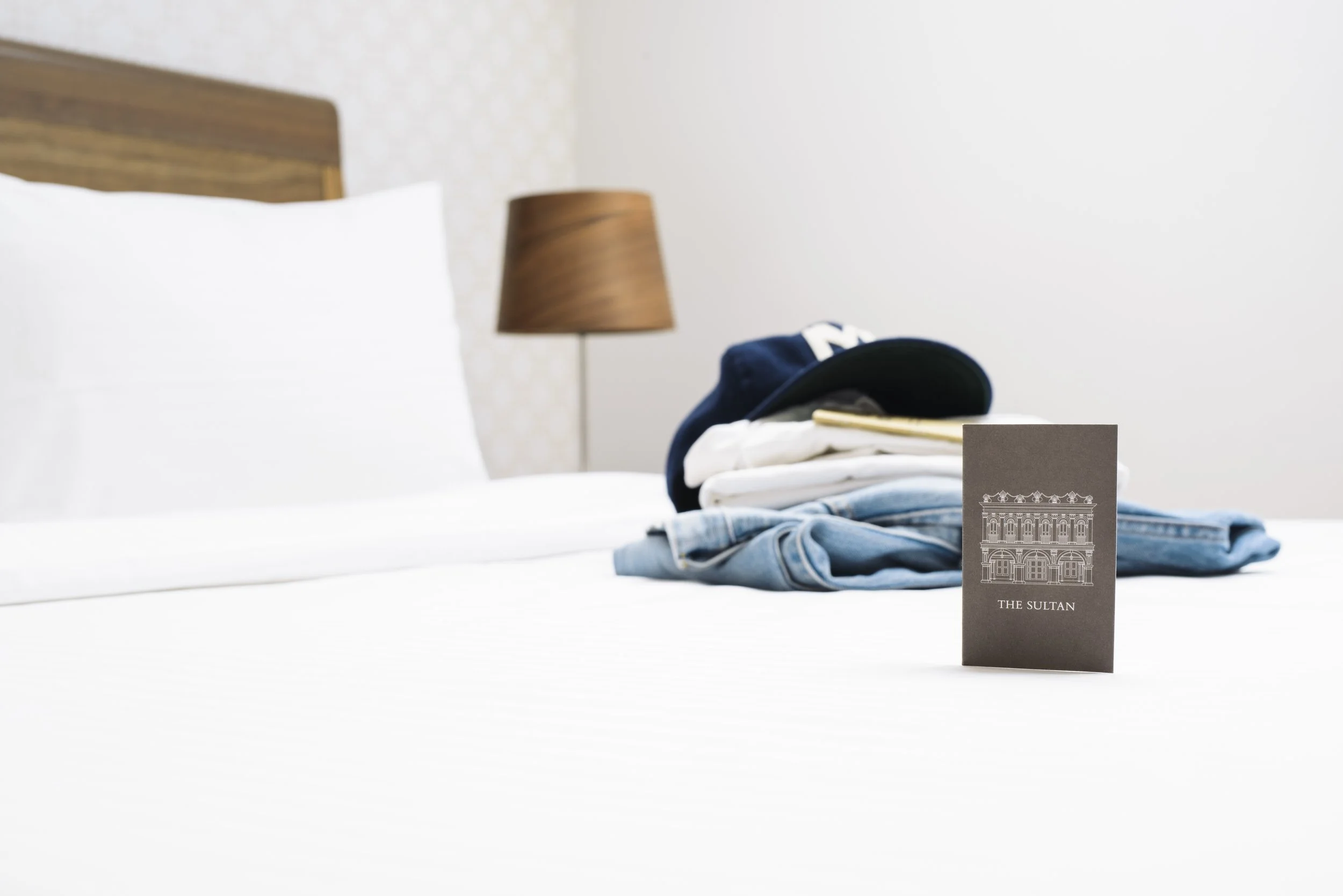 A hotel room with a bed, a wooden headboard, a bedside lamp, and clothes and a cap on the bed, with a card labeled 'The Sultan' in front.
