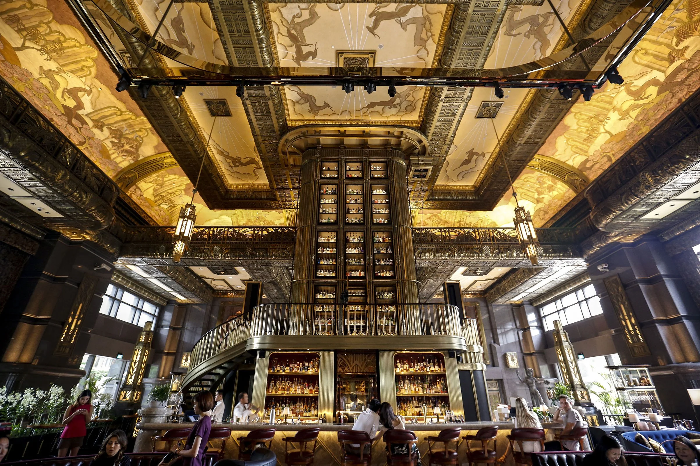 Elegant bar with a tall central liquor display, ornate ceiling with gold accents and mural artwork, patrons sitting at bar and tables, large windows, and decorative lighting.