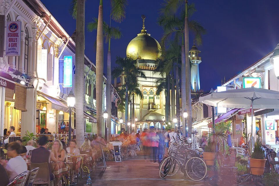 Night scene of a lively outdoor shopping and dining area with tables and bicycles, palm trees, and a prominent golden-domed building in the background.