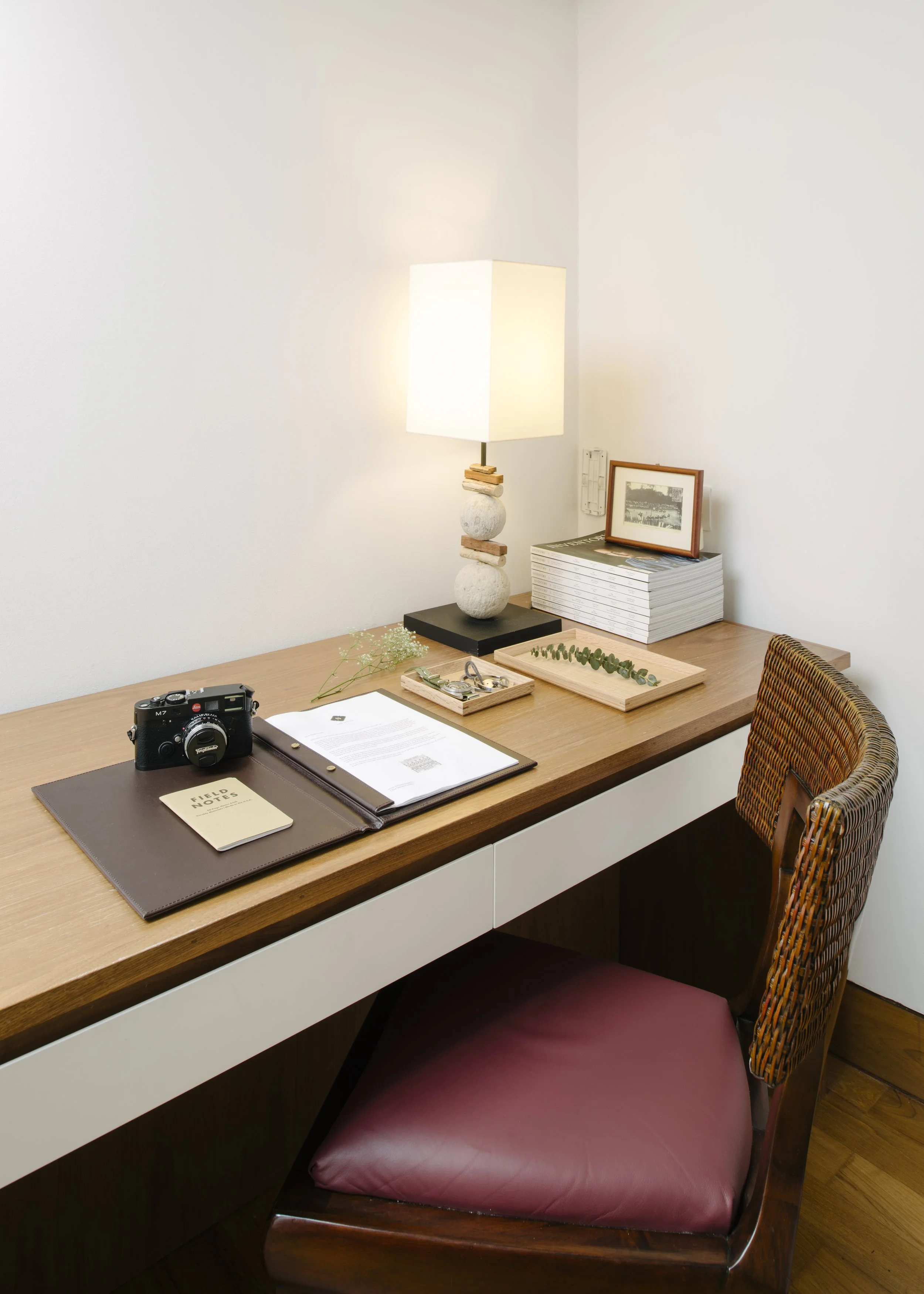 A wooden desk with a brown leather notebook, a black camera, a stack of documents, and a small tray with jewelry. A beige lamp with a square shade, a framed photo, and a large stack of magazines or books are on the desk. Wicker chair with a pink cushion is in front of the desk against white walls.