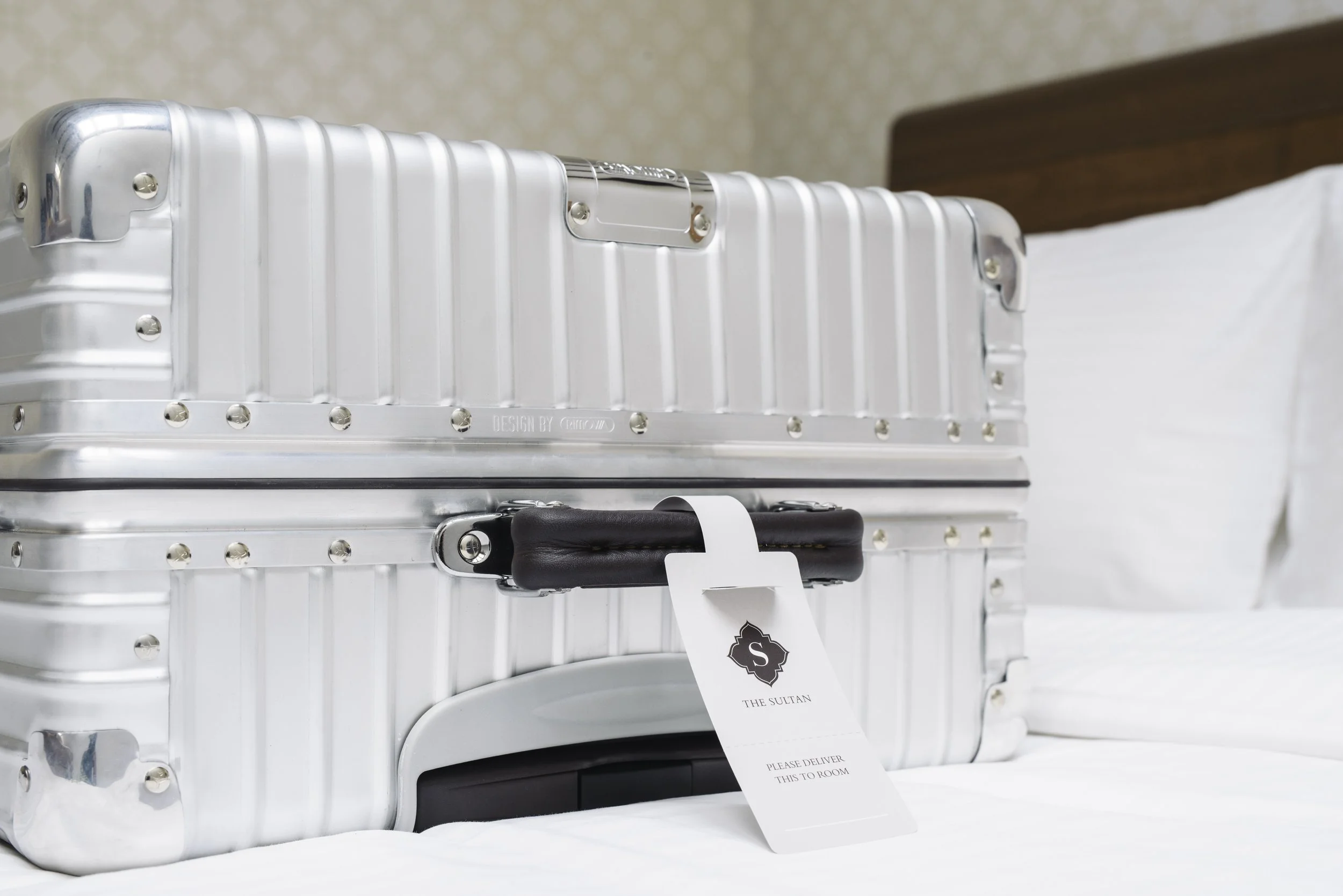 Silver suitcase with black handle and white luggage tag on a bed with white sheets and pillows.