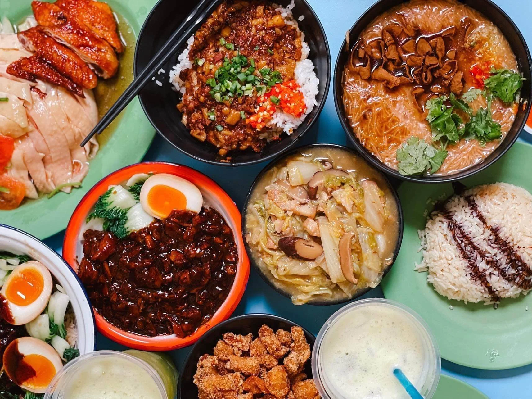 An assortment of Asian dishes including rice, noodles, fried chicken, braised pork, vegetable stir-fry, boiled eggs, and drinks