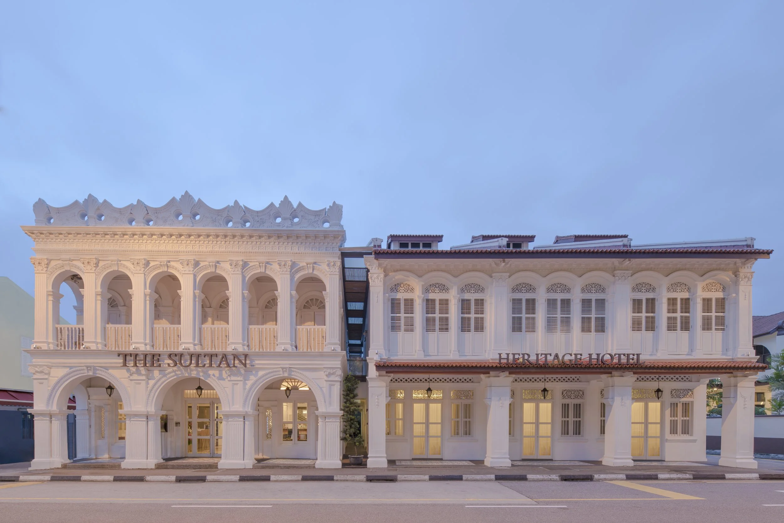 Two white historic buildings with colonial architectural style, labeled 'The Sultan' and 'Heritage Hotel', situated along a street with a sidewalk and a road in front, during the evening with blue sky overhead.