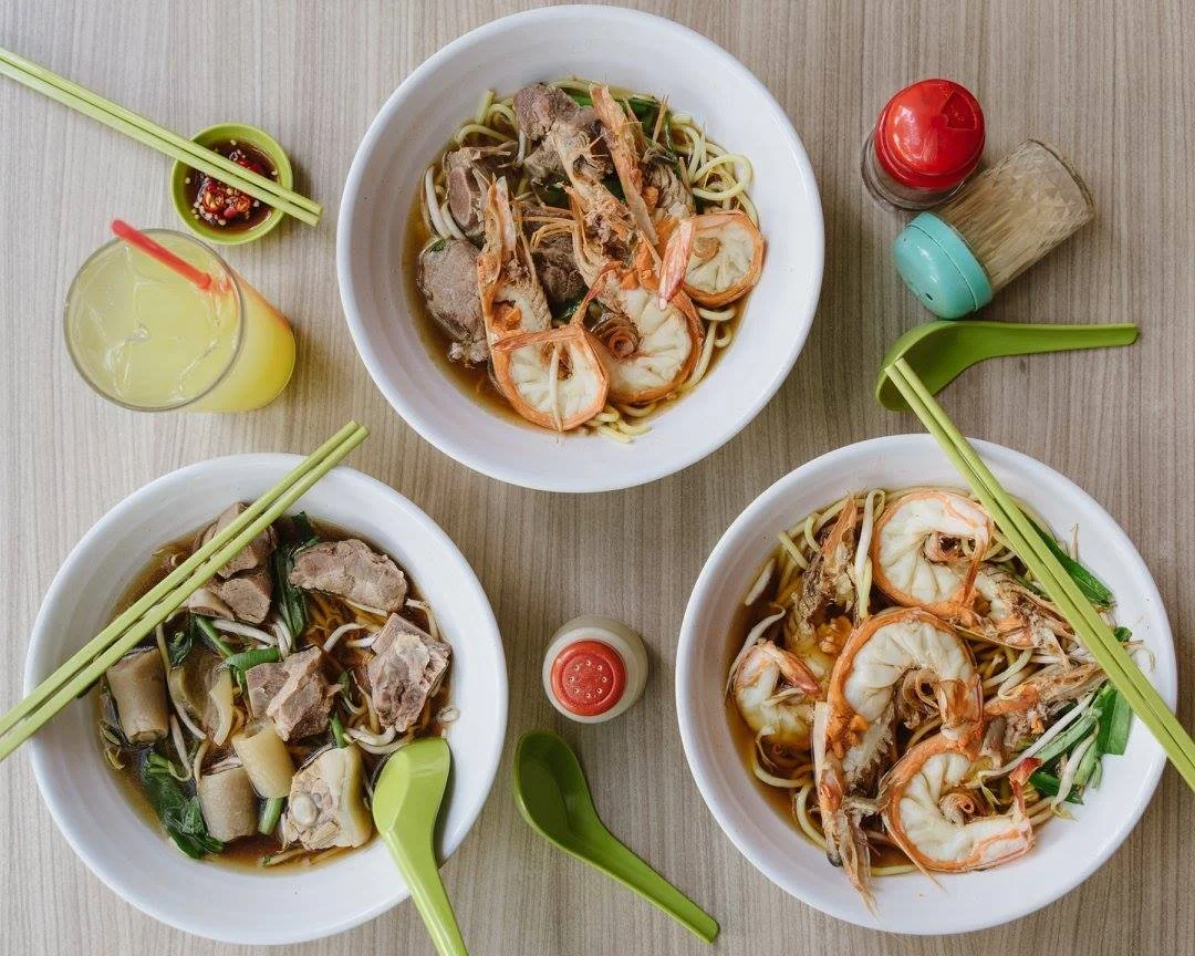 Three bowls of noodle soup with shrimp and meat, soy sauce, and green vegetables, with condiments and drinks on a wooden table.