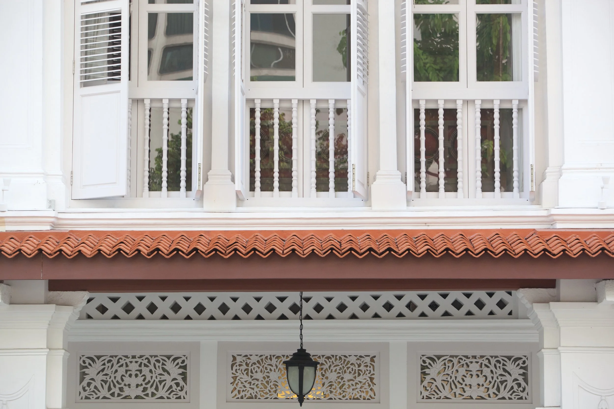 A front view of a white house with a balcony featuring white double doors, decorative balusters, a terracotta tile roof, and a hanging lantern light in front of intricate white latticework.