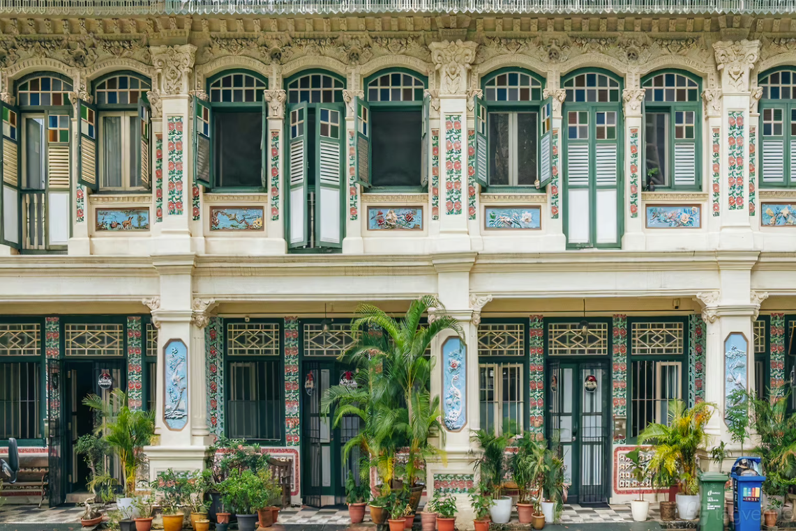 Two-story ornate building with decorative architecture, multiple windows with shutters, and potted plants in front.