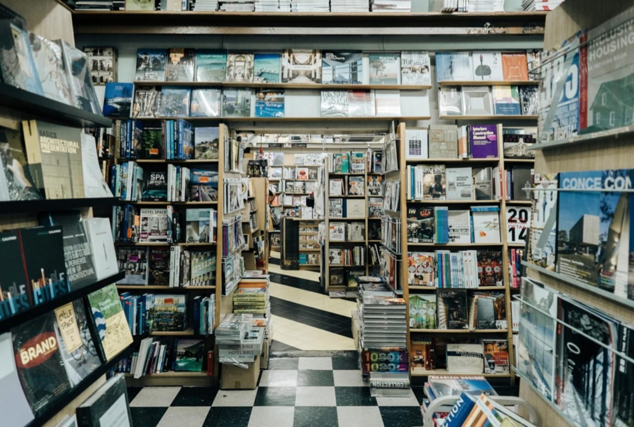 View inside a bookstore with bookshelves filled with magazines and books, black and white checkered floor, and a narrow aisle leading to other sections.