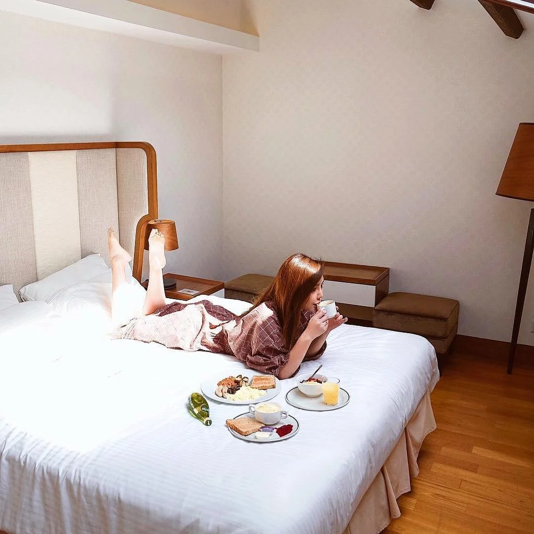 A woman is lying on her stomach on a bed, drinking from a cup, with breakfast foods and drinks on the bed in front of her in a hotel room.