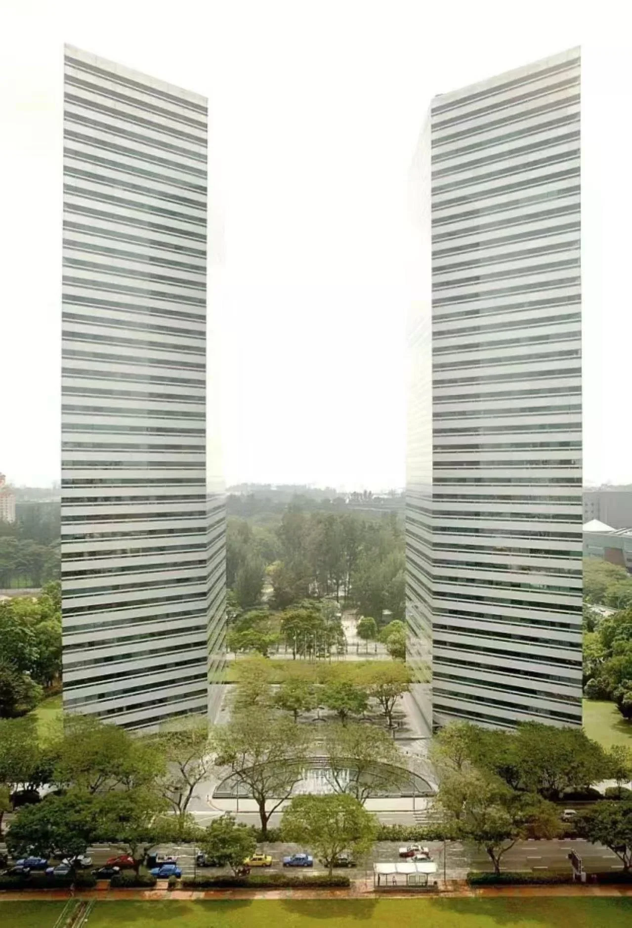 Two modern skyscrapers with glass facades reflecting the sky and clouds, surrounded by greenery and garden paths.