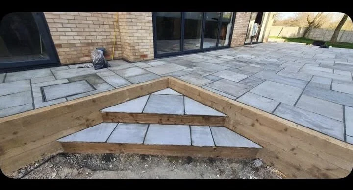 Close-up of a newly built stone and wood staircase leading from a patio to a yard near a brick house with large glass doors.