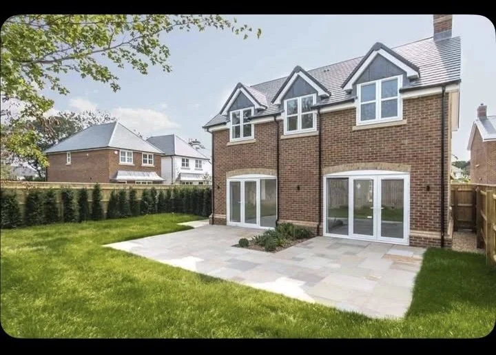 Backyard view of a two-story brick house with large windows and sliding glass doors, surrounded by a green lawn, paved patio, and neighboring houses in the background.