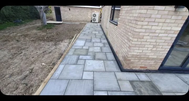 Newly installed stone patio or pathway runs along the outside of a brick house, with an area of bare dirt on one side and an air conditioning unit in the background.