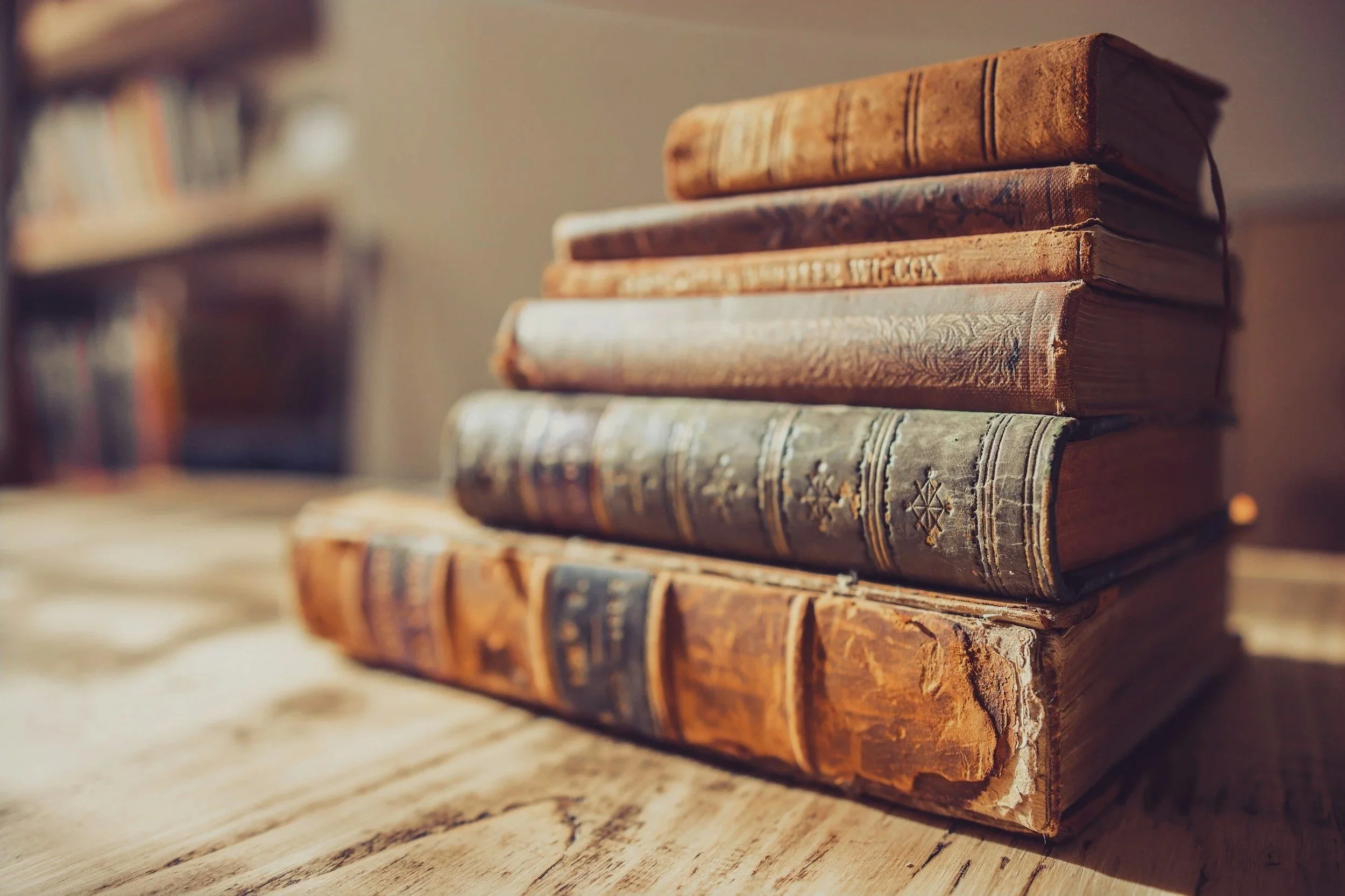 Fünf alte, gebundene Bücher stapeln sich auf einem Holztisch, mit einem Buchregal im Hintergrund.