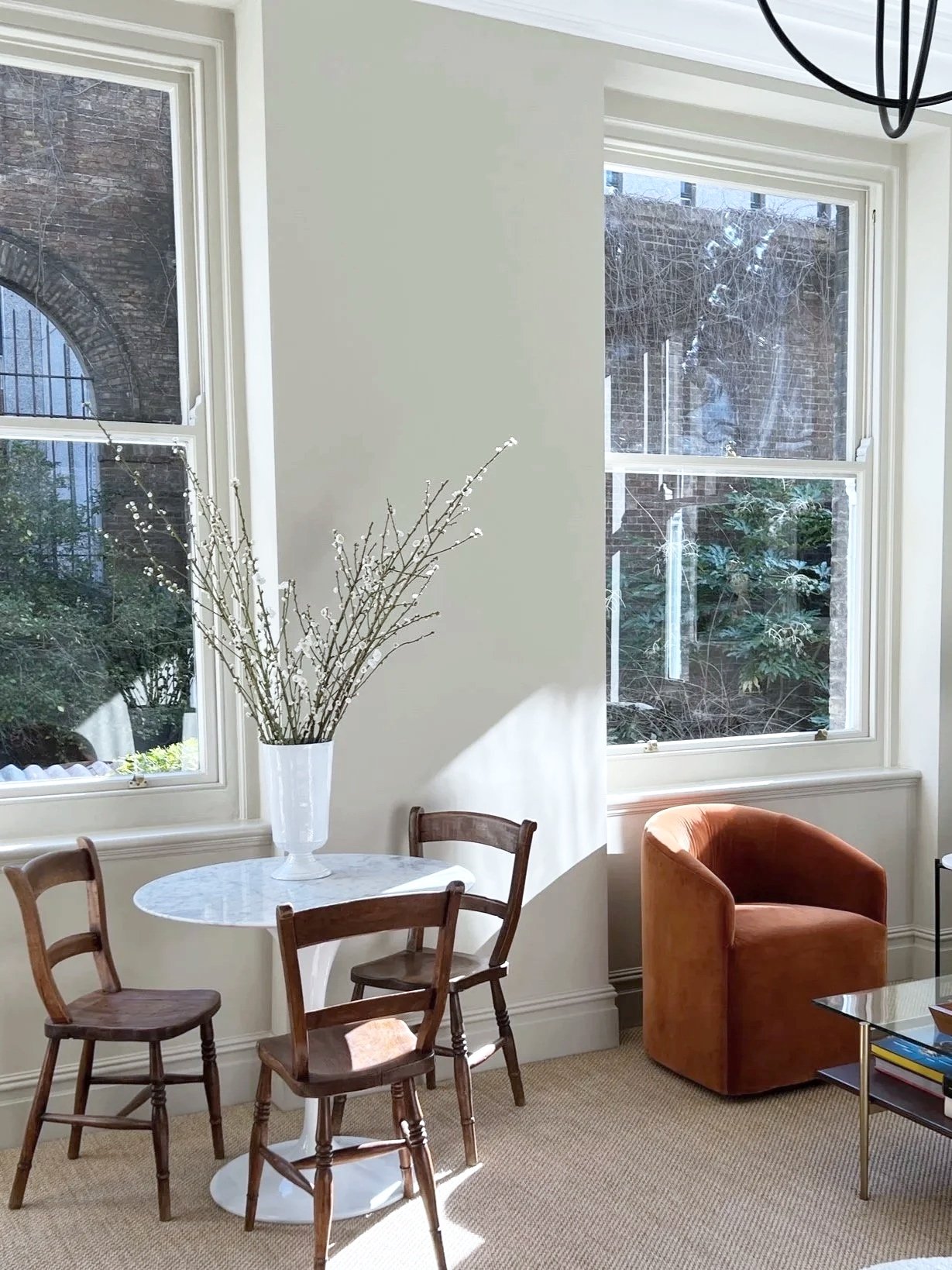 A corner of a living room with two large windows letting in natural light. There is a small round table with a white marble top and three wooden chairs around it. A tall white vase with branches is on the table. An orange armchair is positioned near the windows, and a glass coffee table with books is in front of it.
