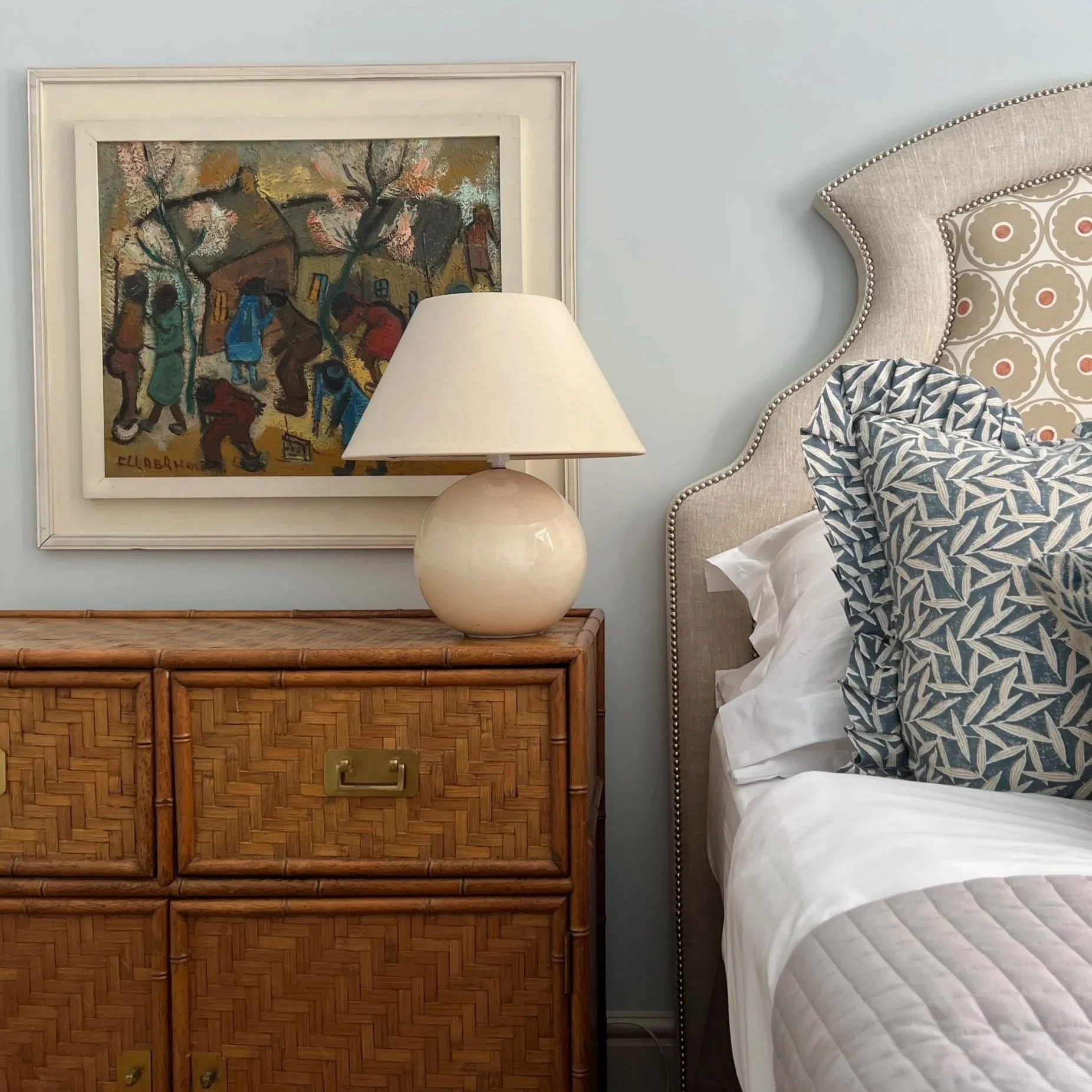 A bedroom with a rattan dresser, a lamp on top, a painting on the wall, and part of a bed with decorative pillows and an upholstered headboard.