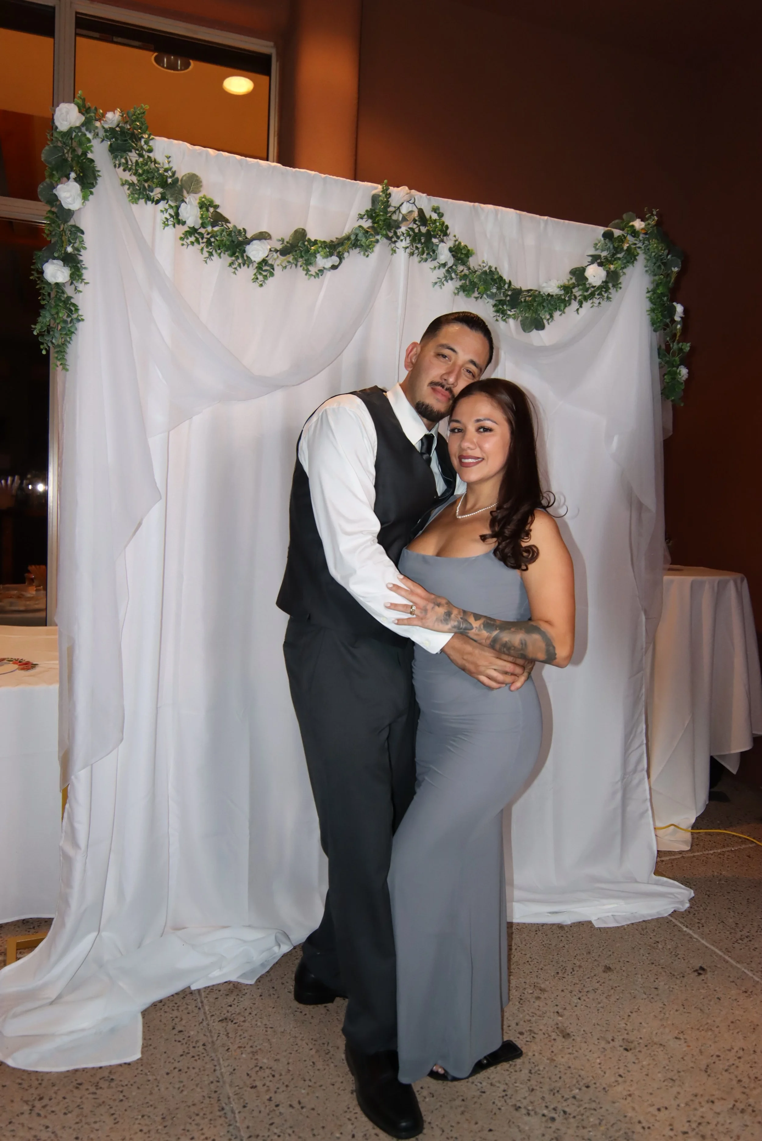 A couple dressed in semi-formal attire standing in front of a white fabric backdrop decorated with greenery and white roses, at a social event or celebration.