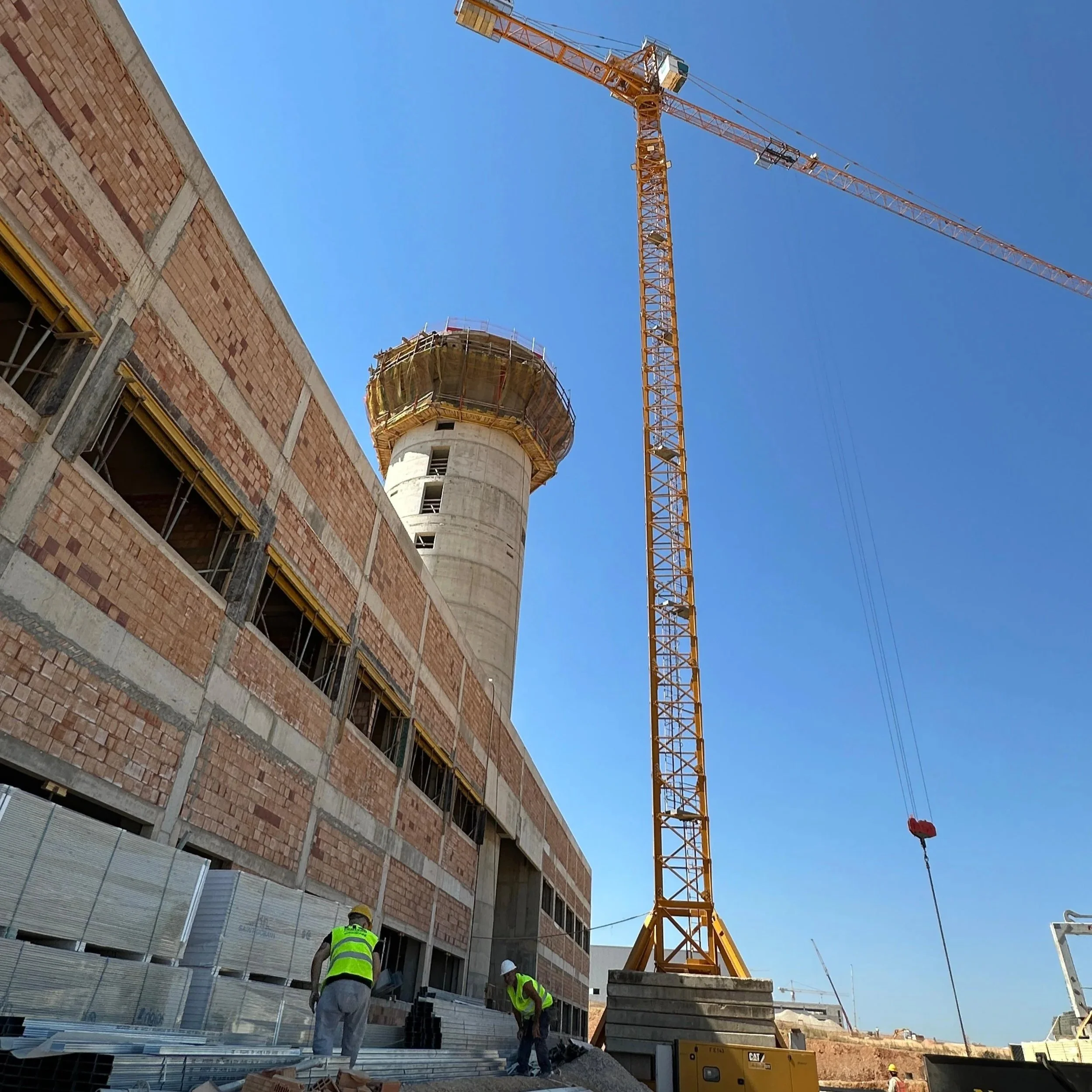 Construction site with workers, a tall crane, and new airport of Heraklion, Crete under construction, with a blue sky in the background.