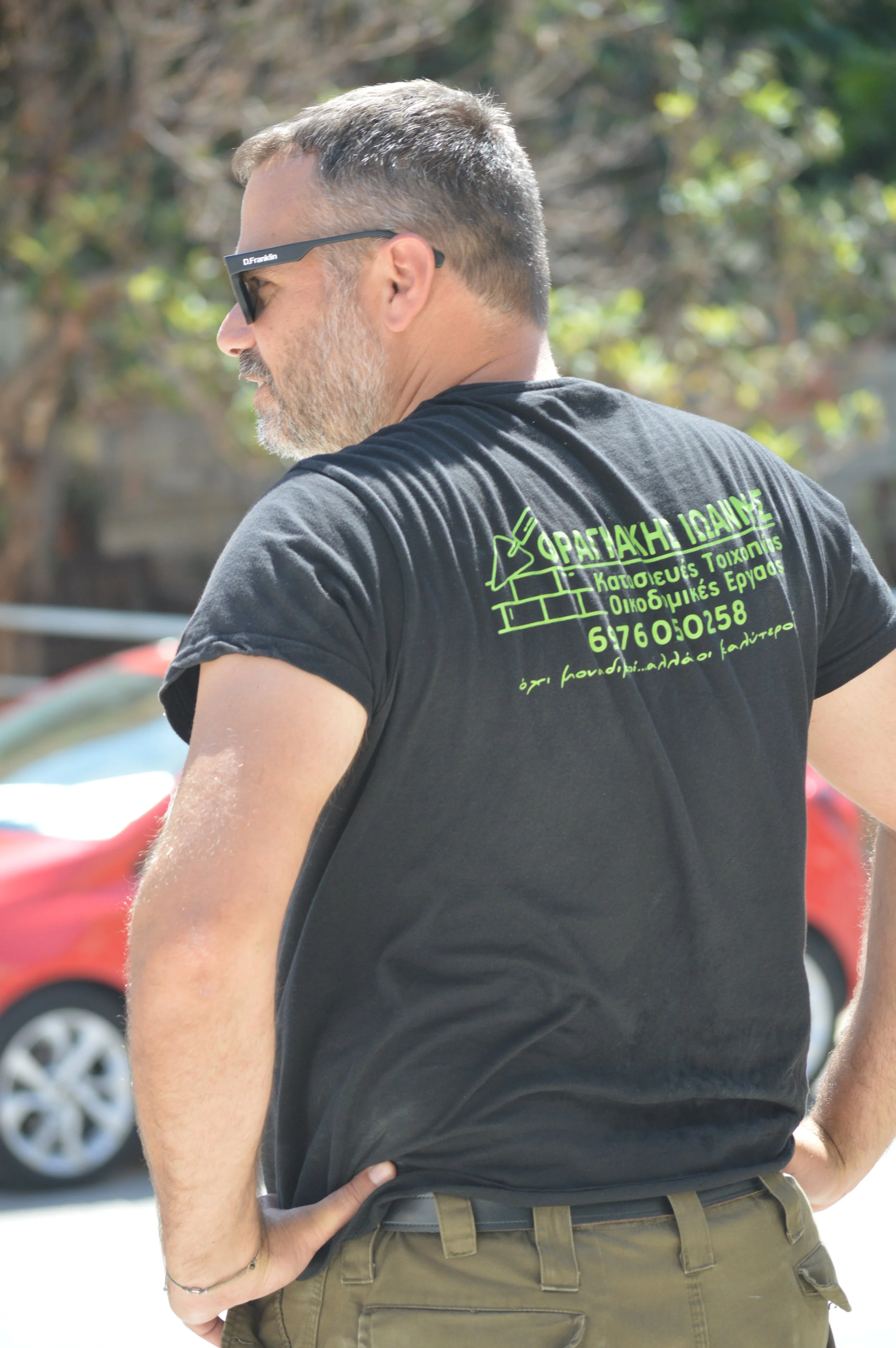A man with short hair, a beard, wearing sunglasses, a black T-shirt, and khaki pants is standing outdoors. The back of his T-shirt has green Greek writing and a graphic of a brick wall.