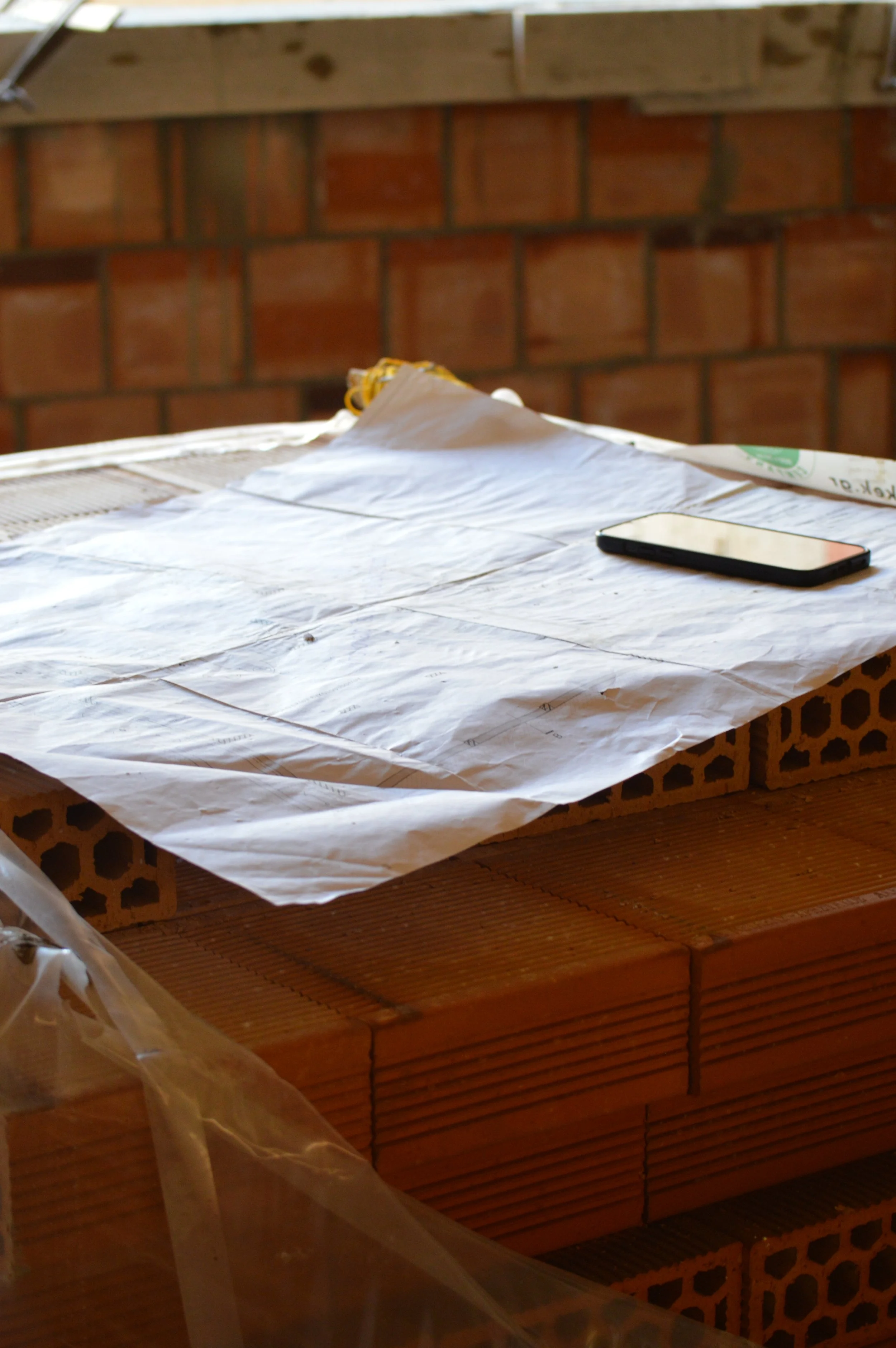 A workspace with architectural plans or blueprints spread out on a brick structure, with a black smartphone placed on top of the papers.