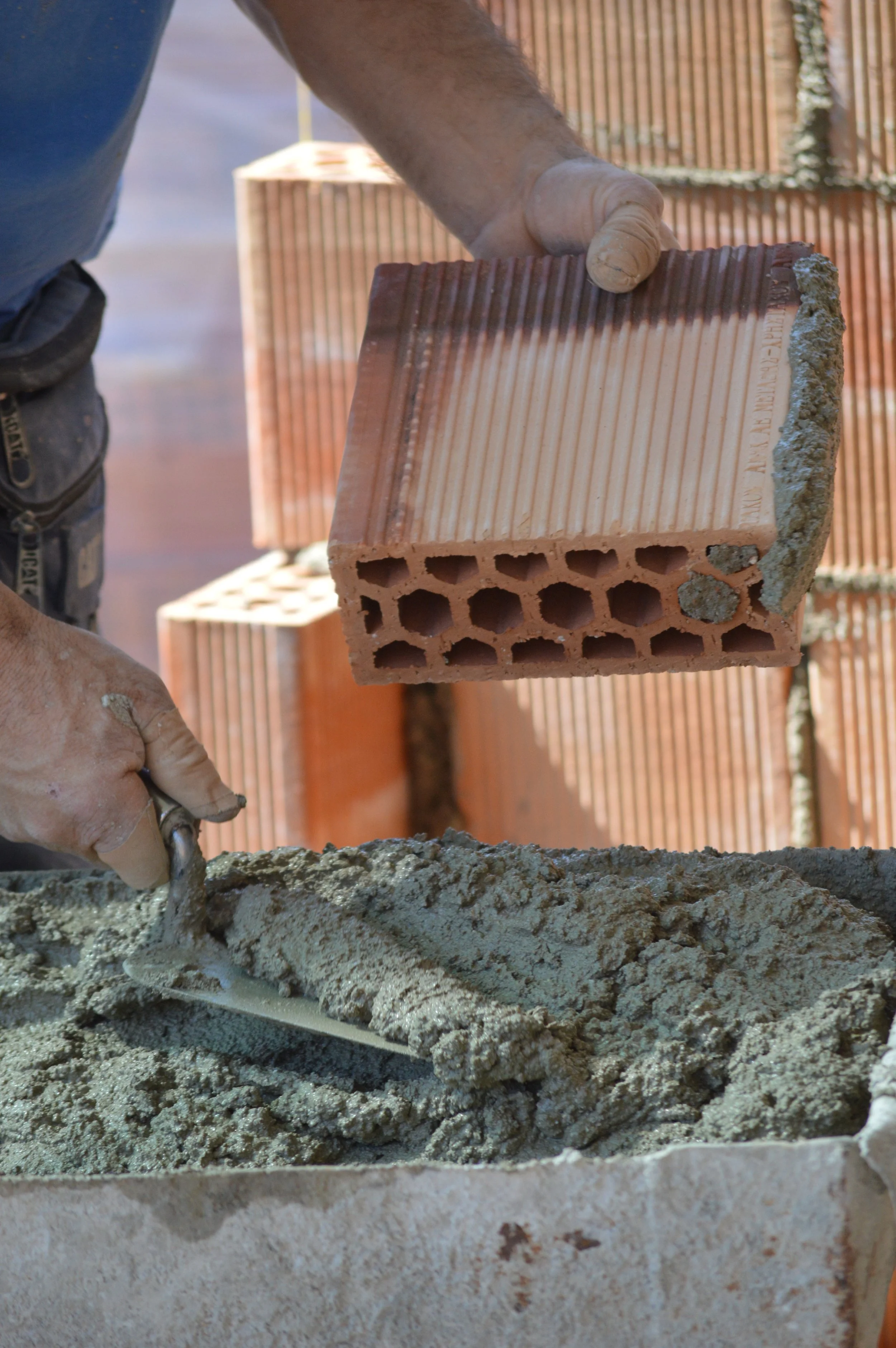 A construction worker is holding a brick while mixing cement with a trowel at a construction site with several bricks in the background.