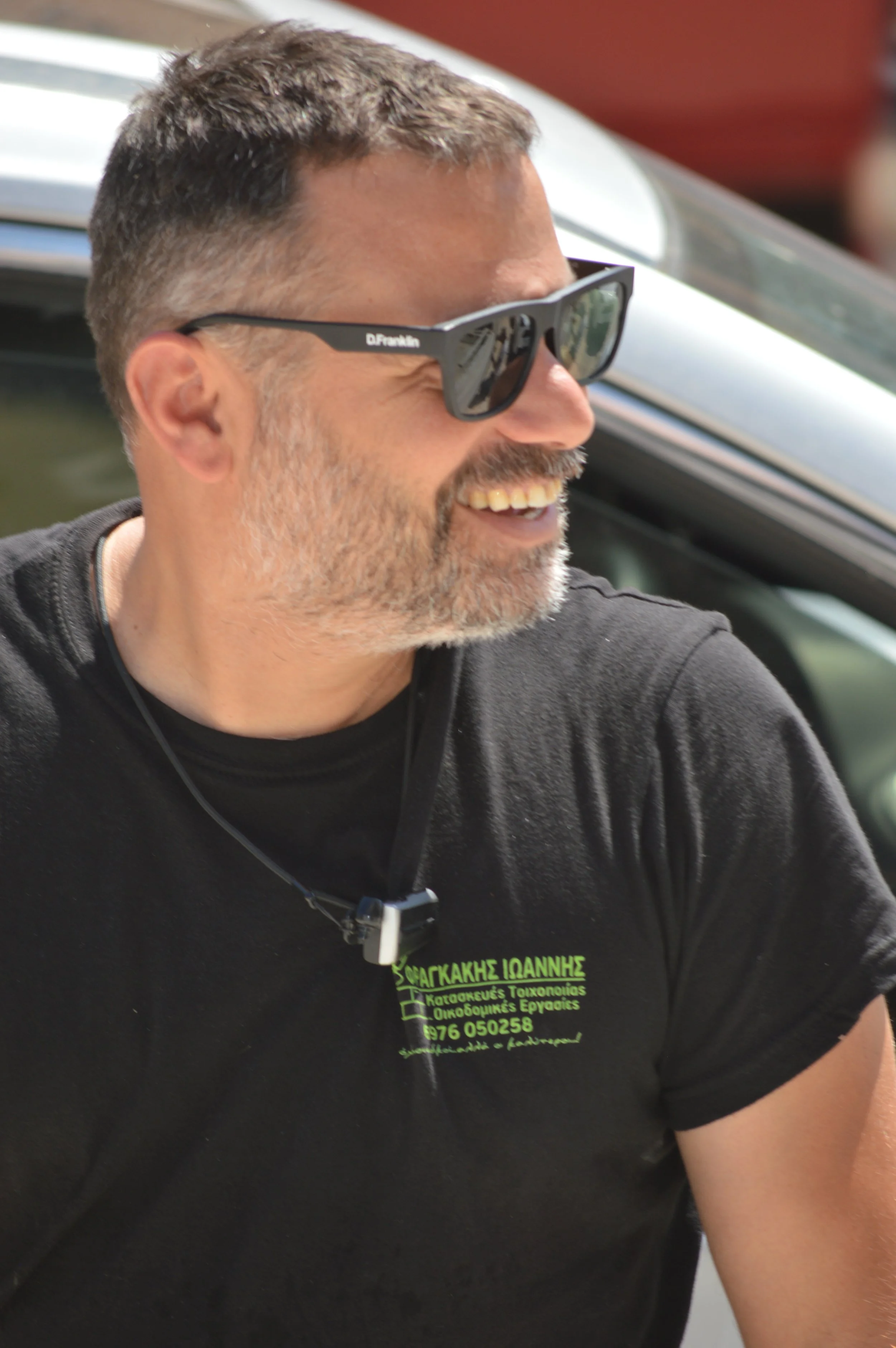 A man with short hair, sunglasses, and a beard, smiling and wearing a black t-shirt with Greek writing, outdoors near a vehicle.