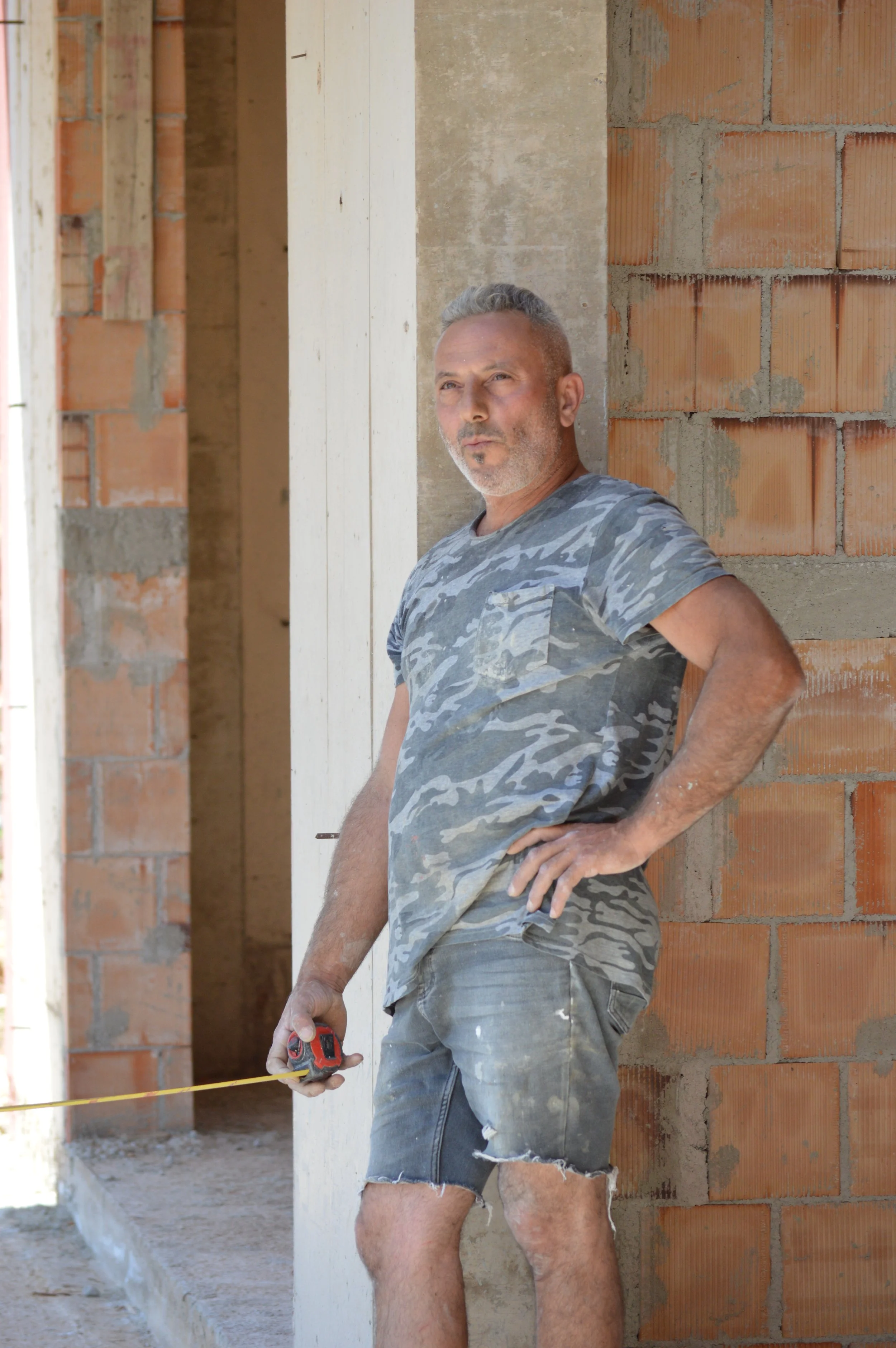 A construction worker, a man, standing outside a building under construction, with one hand on his hip and holding a tape measure in the other. He is wearing a camouflage t-shirt and ripped shorts.