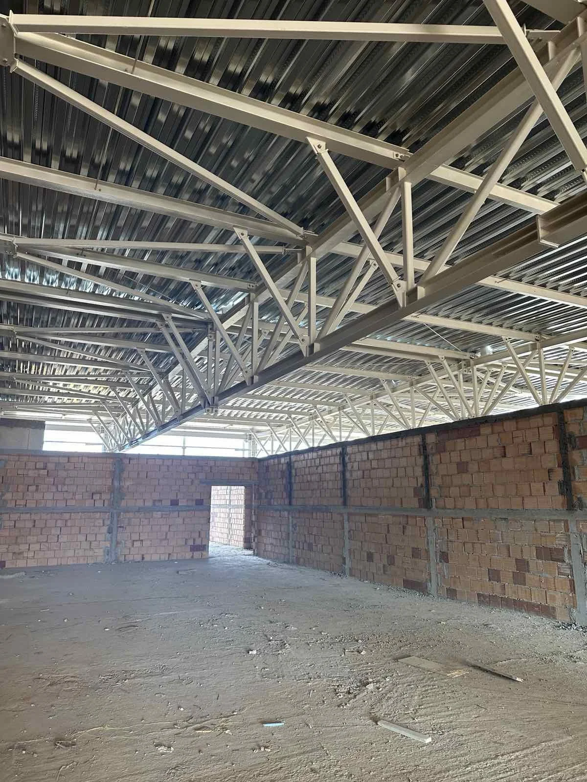 Interior view of a building under construction with brick walls, a dirt floor, and a metal roof ceiling supported by steel trusses.