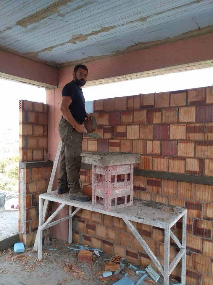 Construction worker standing on a temporary scaffold inside a building under construction, with exposed brick walls and a ceiling with peeling paint.