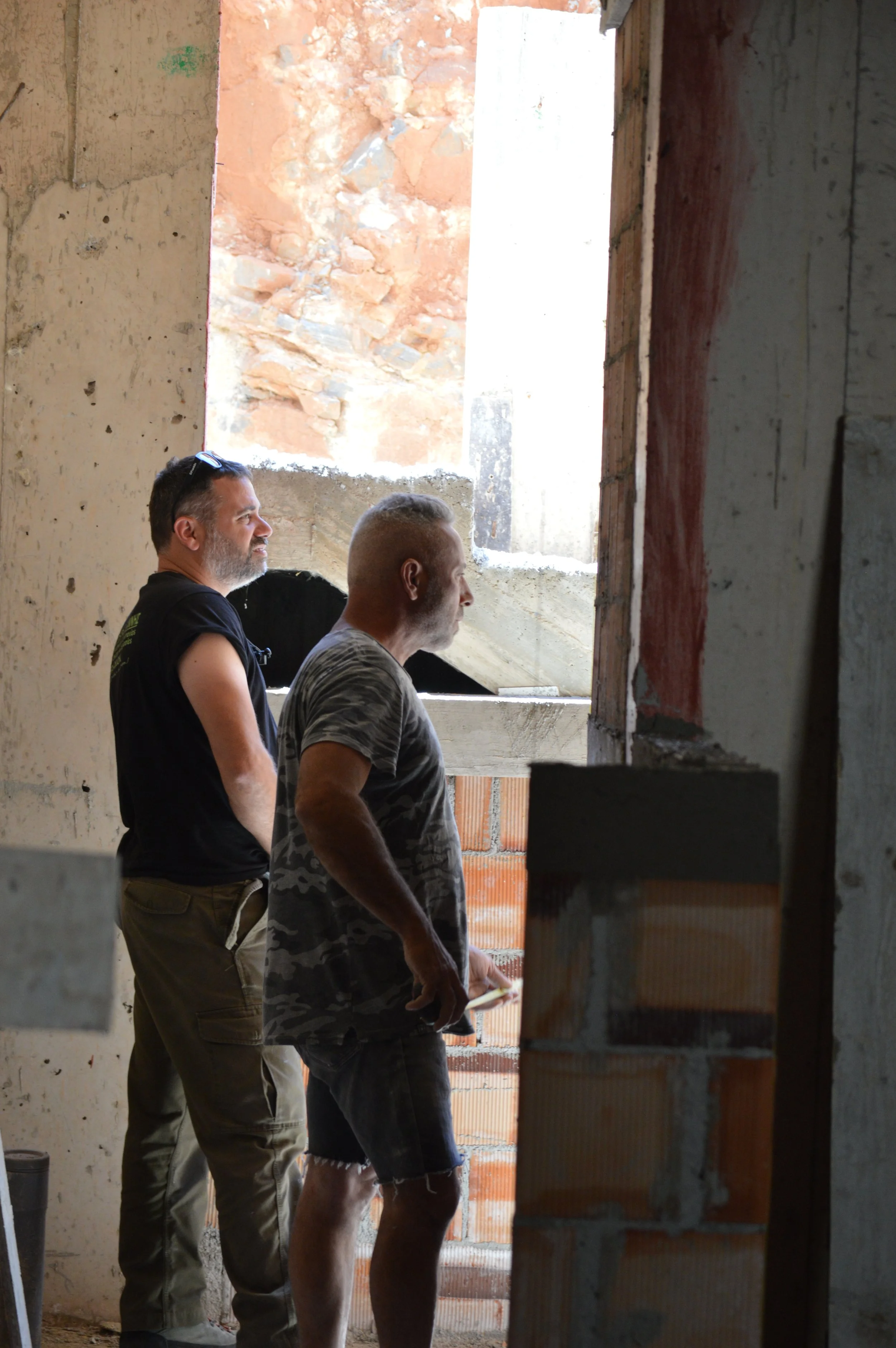 Two men stand inside an unfinished building, looking out a large open window. The walls are made of exposed brick and concrete, and there is construction debris on the floor.