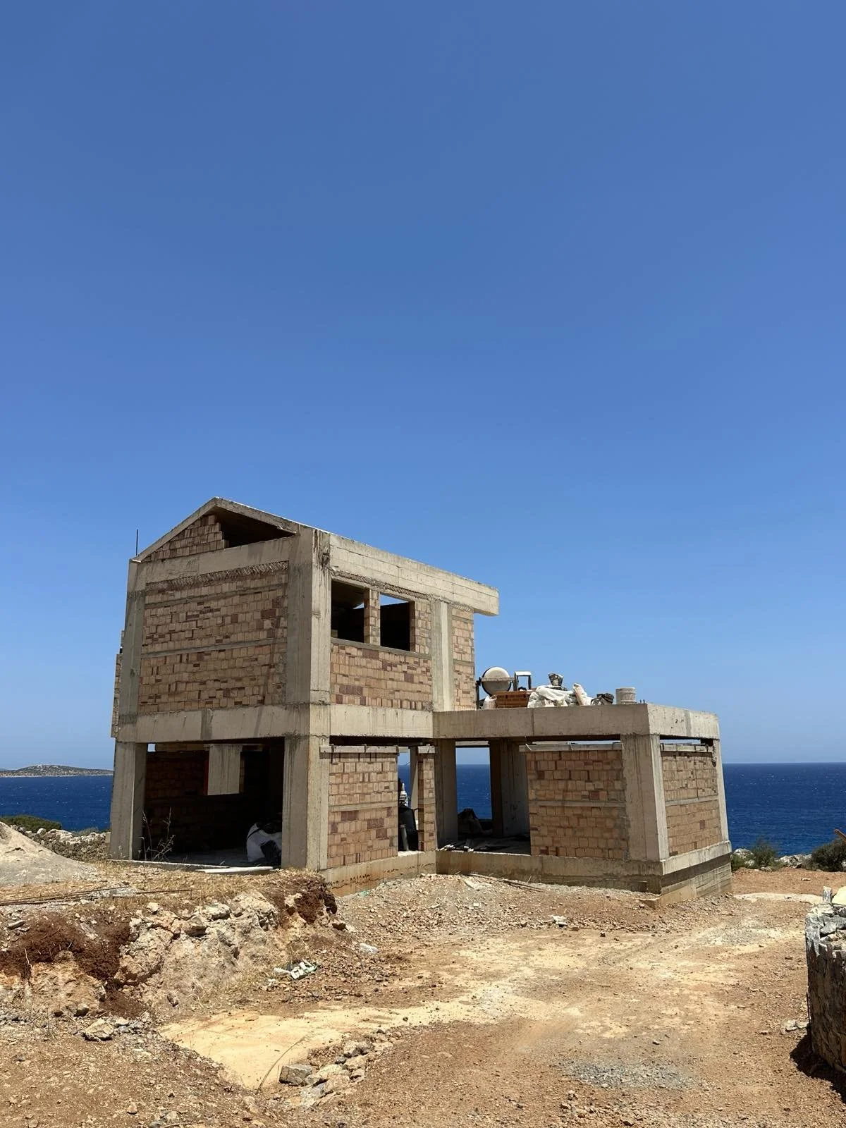 Under construction two-story house with brick walls and concrete frames on a dirt lot near the ocean, clear blue sky overhead.