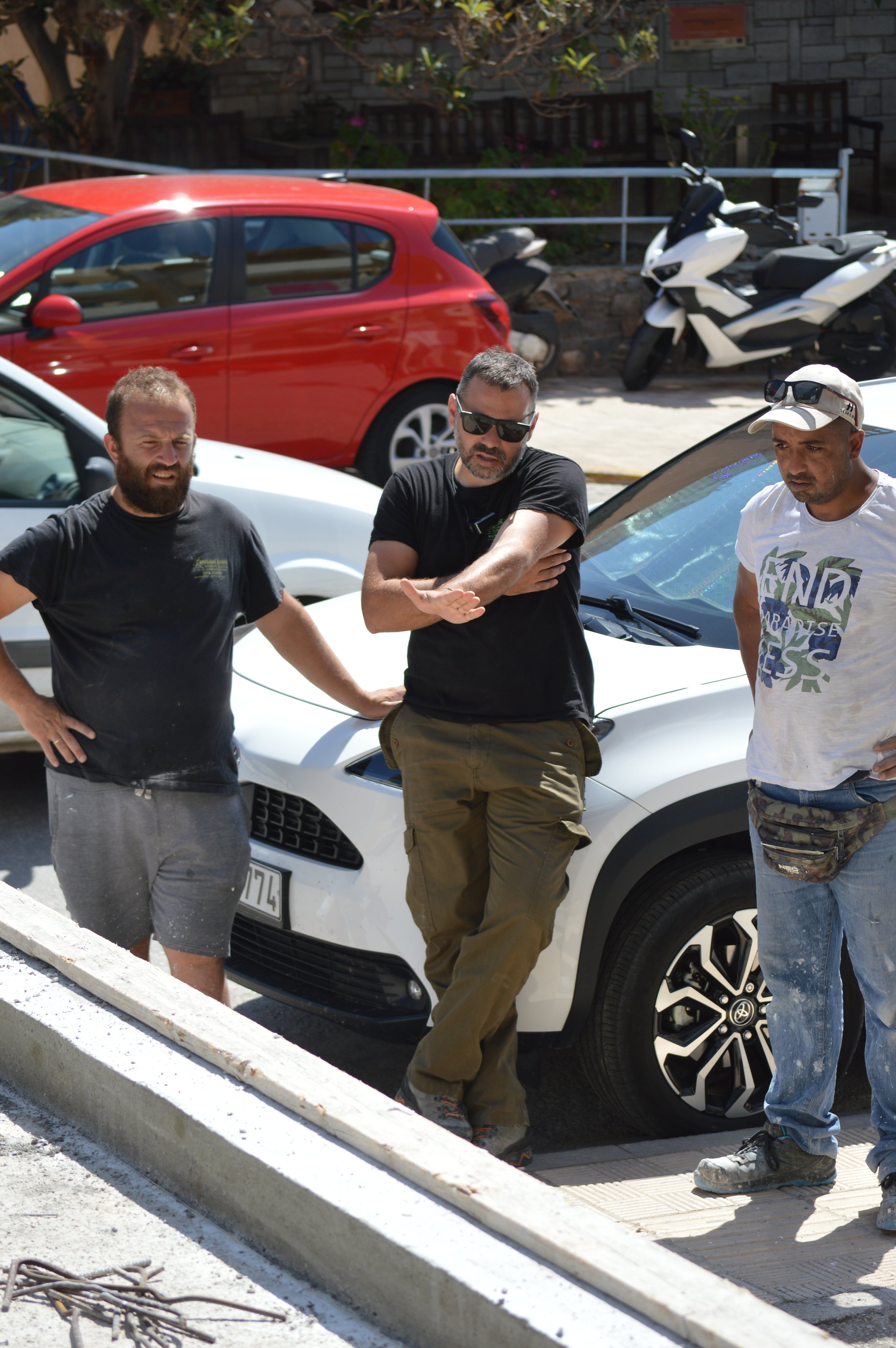 Three men standing and discussing in front of a white car parked on a street, with other cars and scooters in the background.