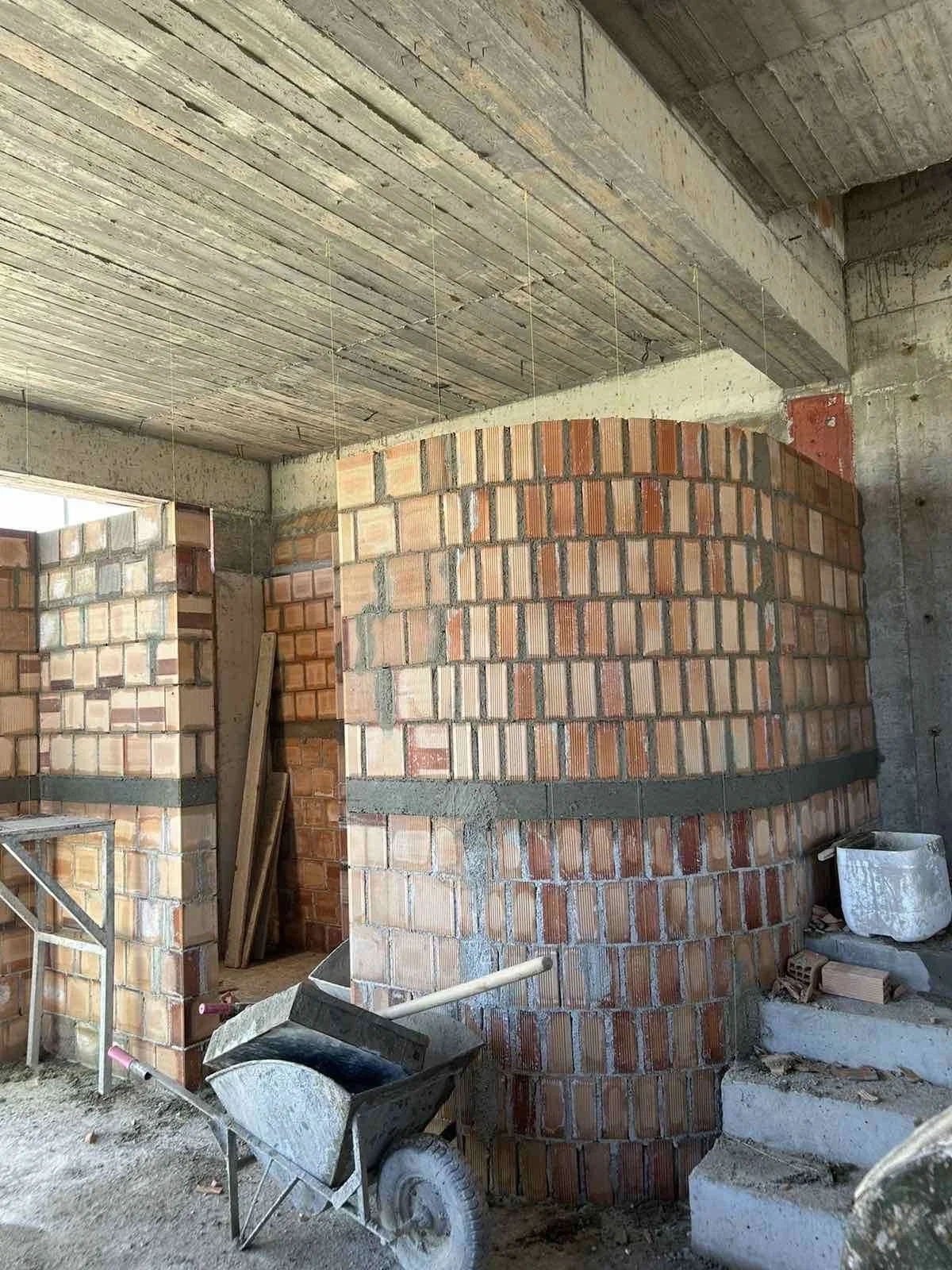 Interior view of a construction site showing brick walls, concrete ceiling, a wheelbarrow, and construction materials.