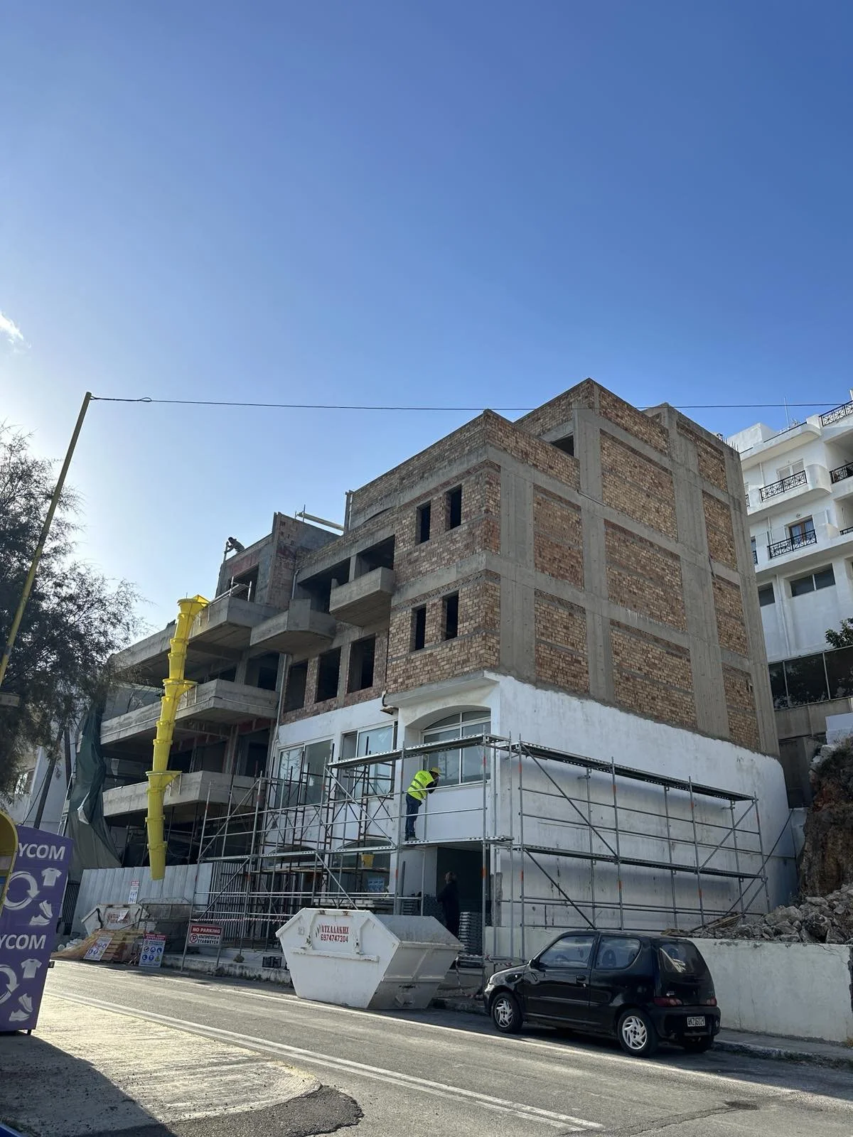 Building under construction with scaffolding and workers on site, a yellow construction chute attached to the building, and a black car parked in front, under a clear blue sky.