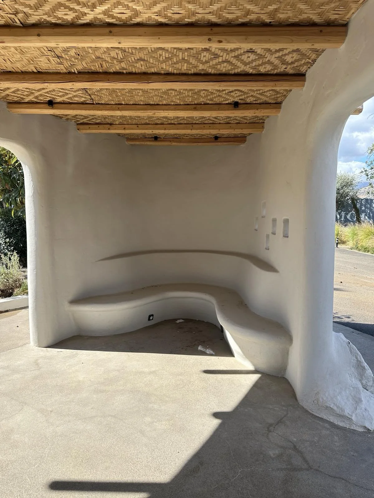 Modern outdoor bus stop shelter with curved white concrete walls, built-in bench, and a wooden slat roof.