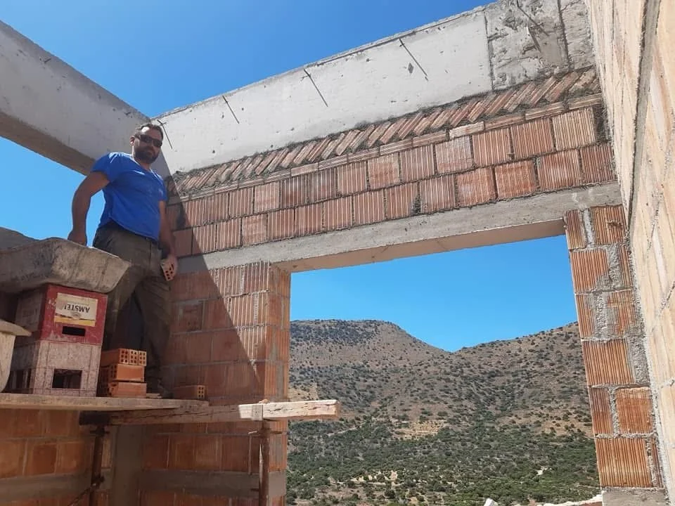 A man wearing a blue shirt and sunglasses standing inside a partially built brick structure with mountains and a clear blue sky in the background.