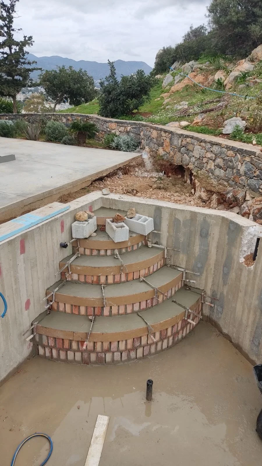 Construction site showing a partially built staircase made of bricks and concrete inside a foundation with soil and rocks around it, and outdoor view of trees, shrubs, and mountains in the background.
