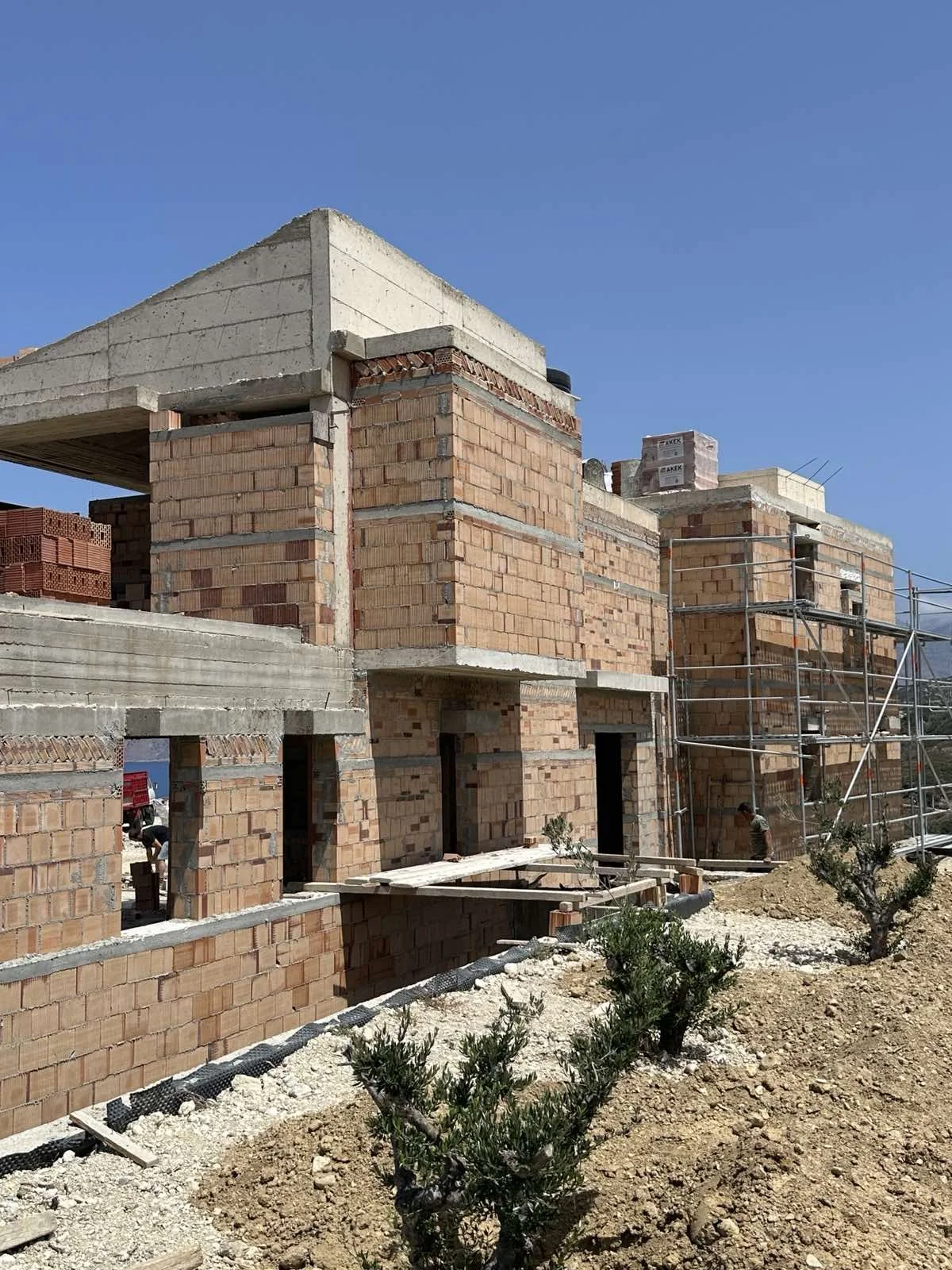 Under construction brick building with scaffolding on one side, small bushes and dirt in foreground, clear blue sky above.